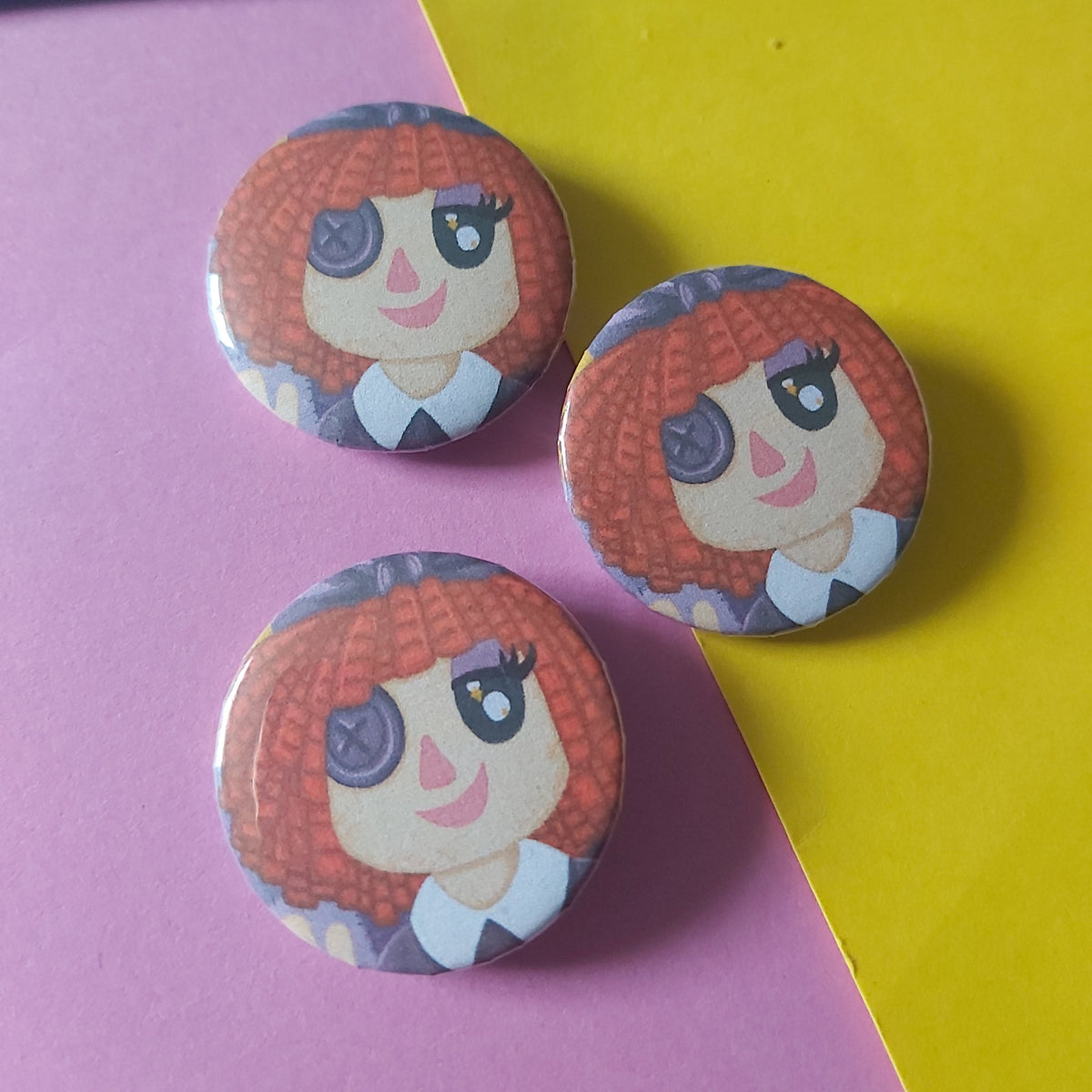 Three round pinback buttons featuring a cartoon girl with red hair and a purple button eye, arranged on a diagonally split pink and yellow background.