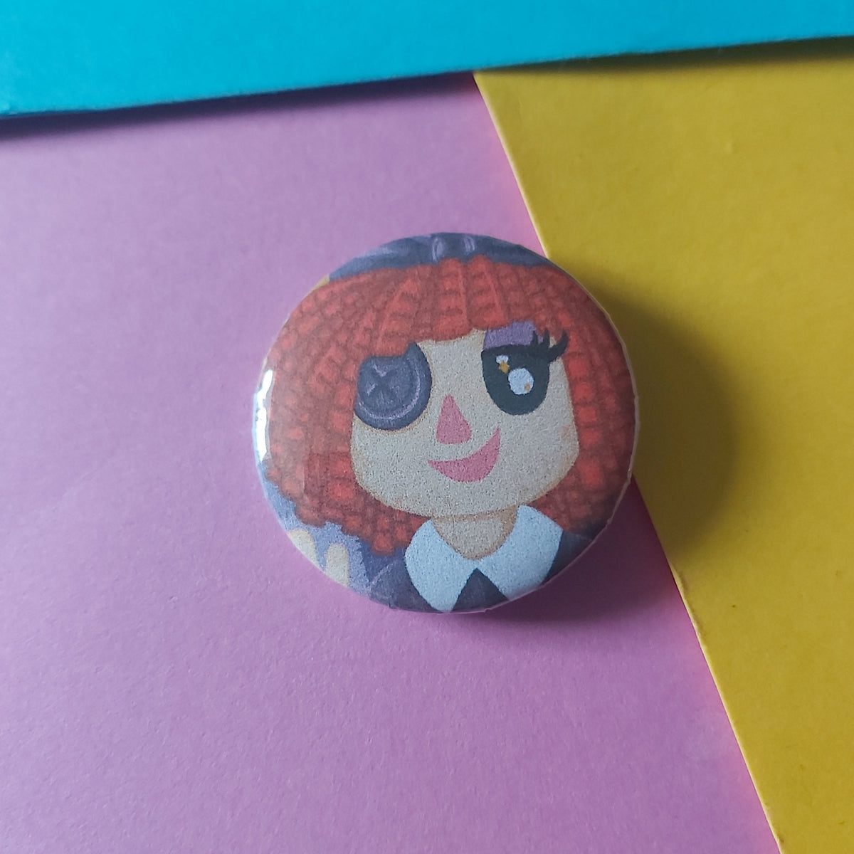 A round pin-back button featuring a cartoon character with red hair and one button eye, placed on a colourful background of purple, yellow, and blue paper.