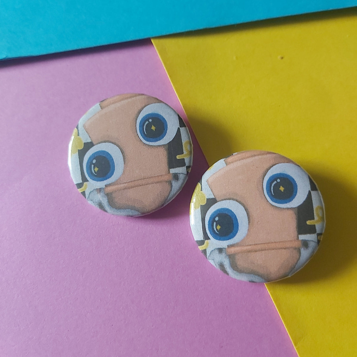 Two round pinback buttons featuring a cartoon chess piece face with large blue eyes and a beige face, displayed on a diagonally split pink, yellow, and blue background.