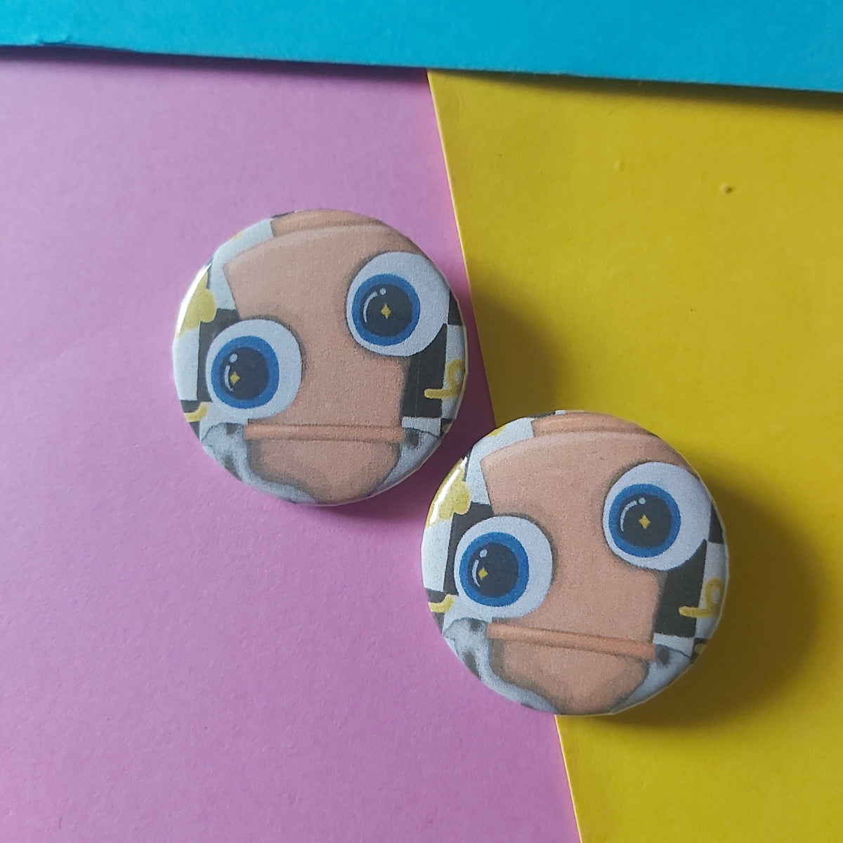 Two round pinback buttons featuring a cartoon chess piece face with large blue eyes, displayed on a split pink and yellow background.