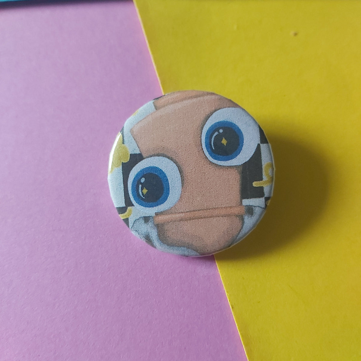 A round pinback button badge featuring a cartoon chess piece face with large blue eyes and a tan head, placed on a diagonally split pink and yellow background.