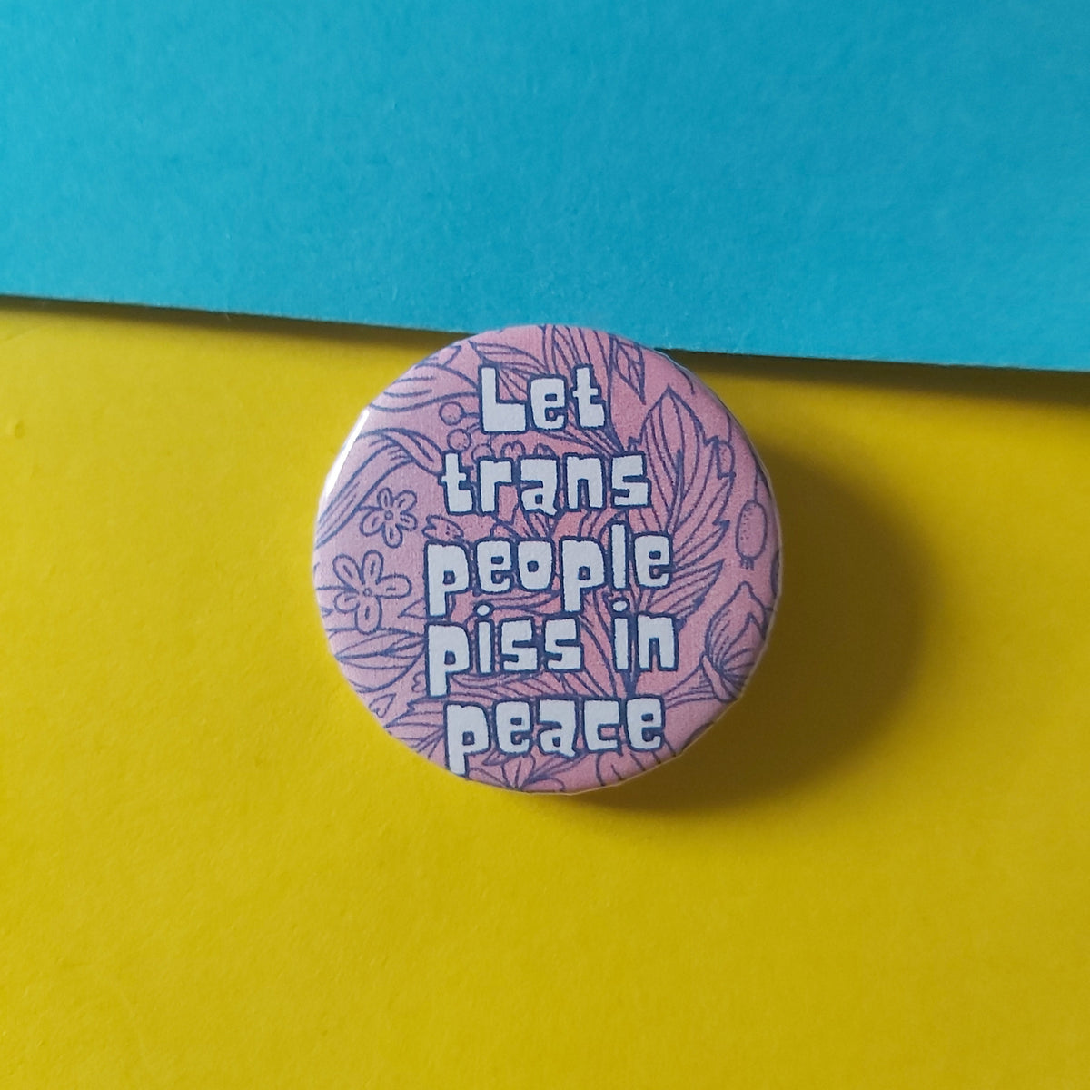 A round pinback button badge featuring the text 'Let trans people piss in peace' in light blue lettering over a lavender floral pattern, displayed on a split yellow and teal background.