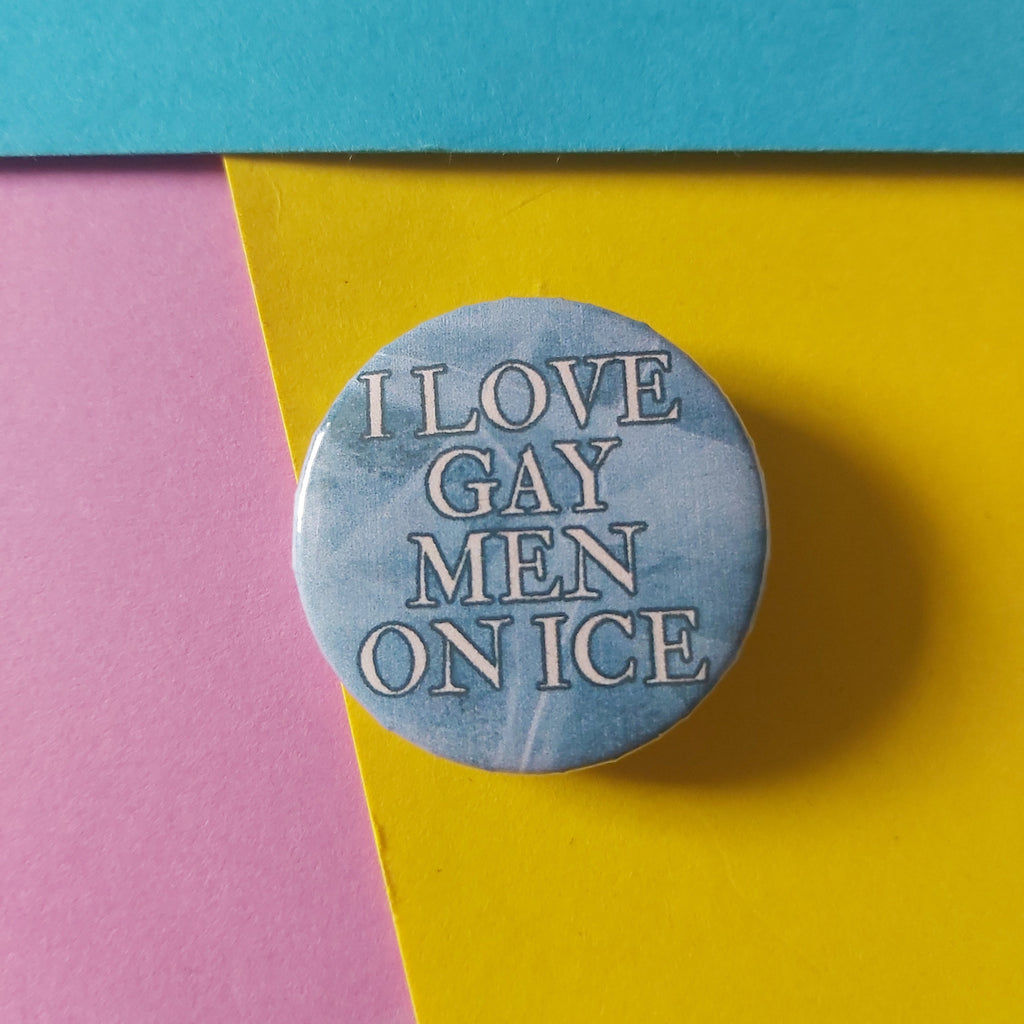 A round pin-back button badge with a light blue icy background and the text 'I LOVE GAY MEN ON ICE' in blue outlined lettering, photographed on a colourful background split diagonally between pink, yellow, and teal paper.