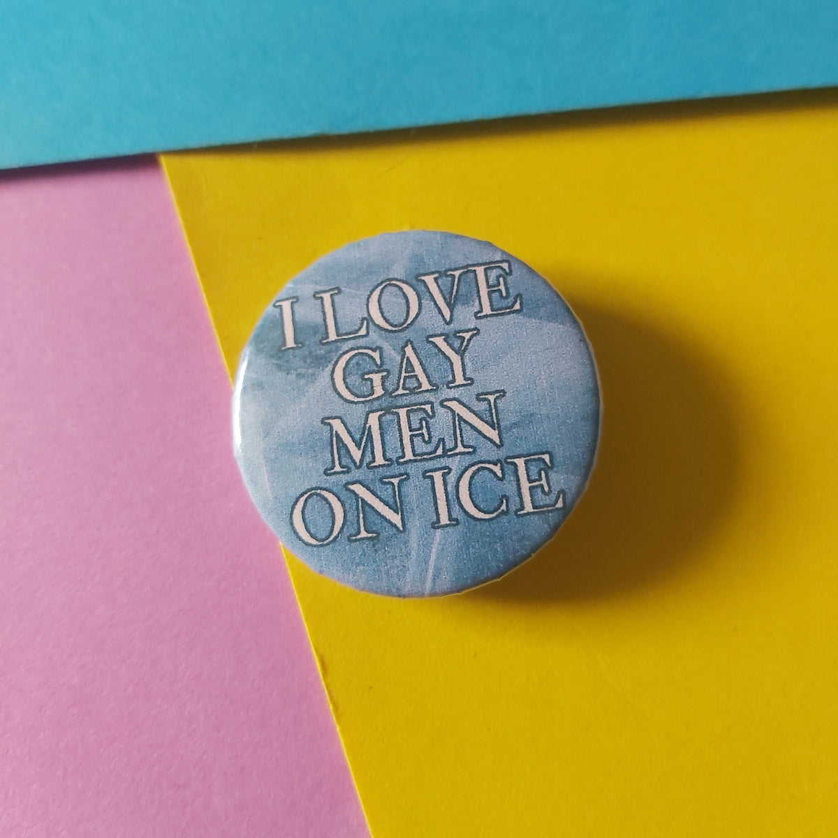 A round pin badge with a light blue and white marble pattern, featuring the text 'I LOVE GAY MEN ON ICE' in white. The pin is displayed on a background of diagonally split pink, yellow, and blue paper.