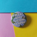 A round button pin with the text 'I'M GAY AND I LISTEN TO KPOP' over a floral background, placed on a colorful background of blue, yellow, and pink paper.
