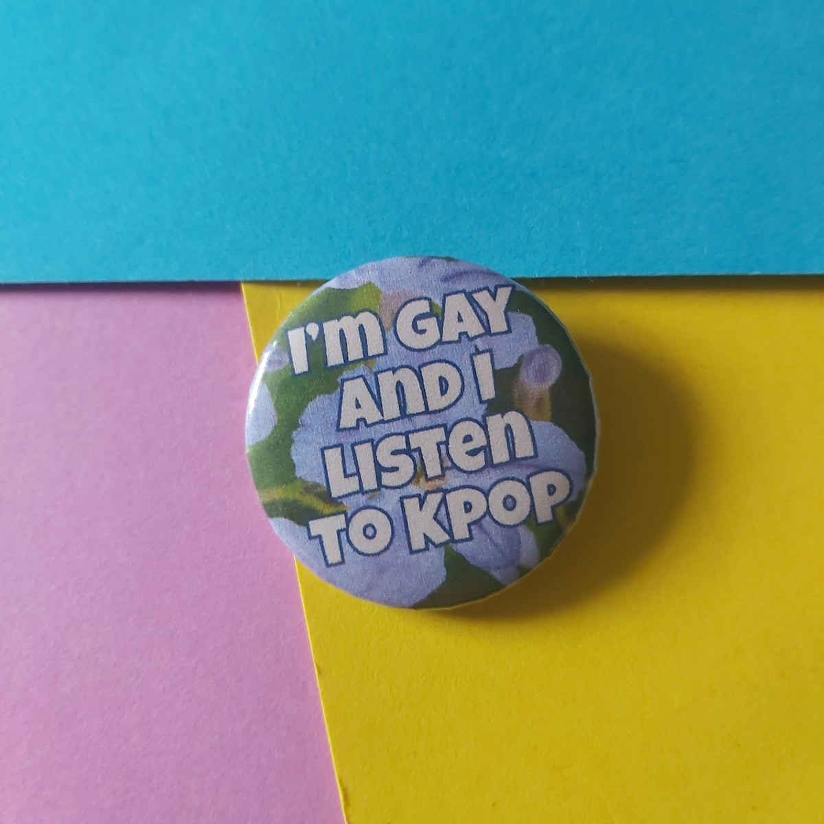 A round button pin with the text 'I'M GAY AND I LISTEN TO KPOP' over a floral background, placed on a colorful background of blue, yellow, and pink paper.