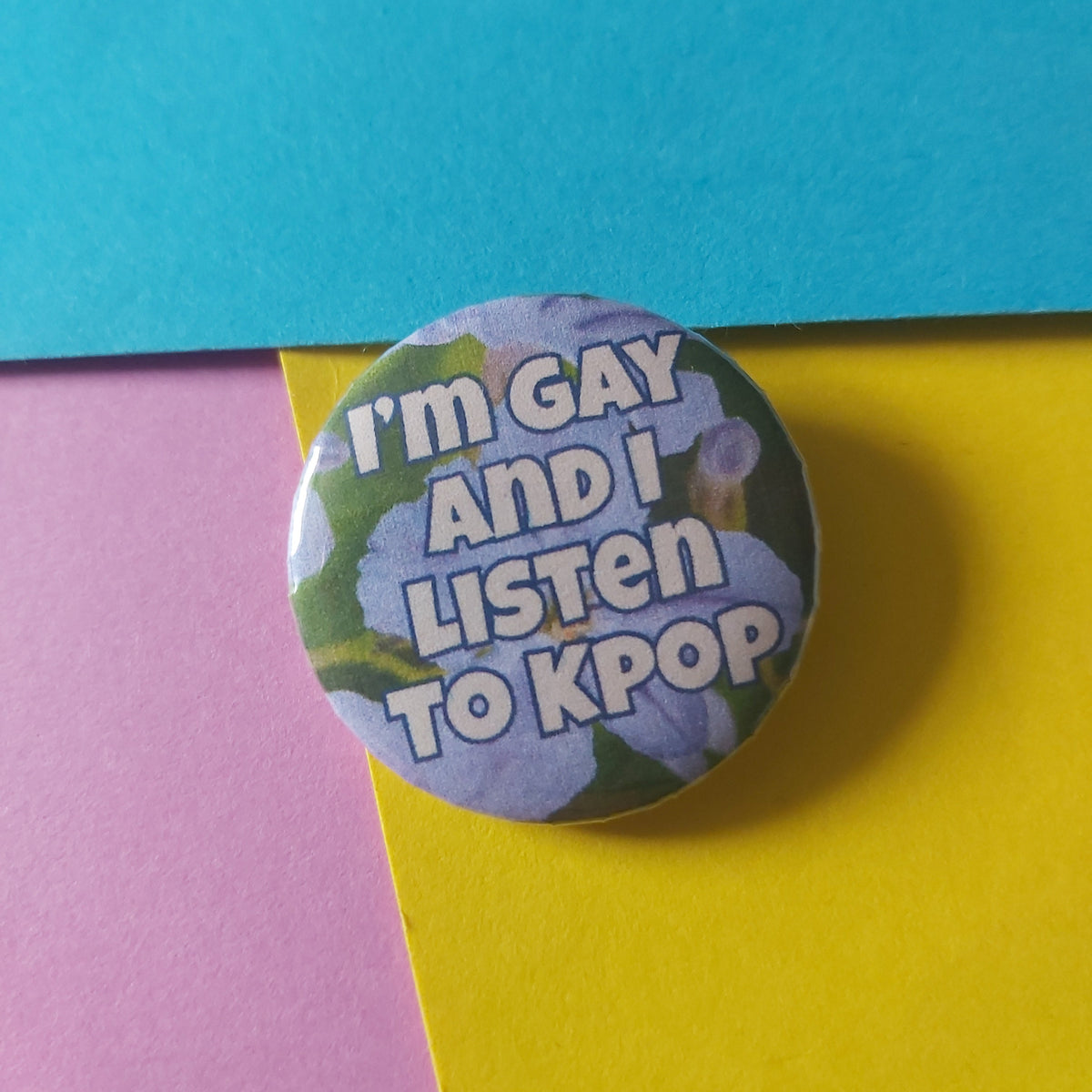 A round button pin with the text 'I'm GAY AND I LISTEN TO KPOP' in white letters over a purple flower background, placed on a colourful background of blue, yellow, and pink paper.