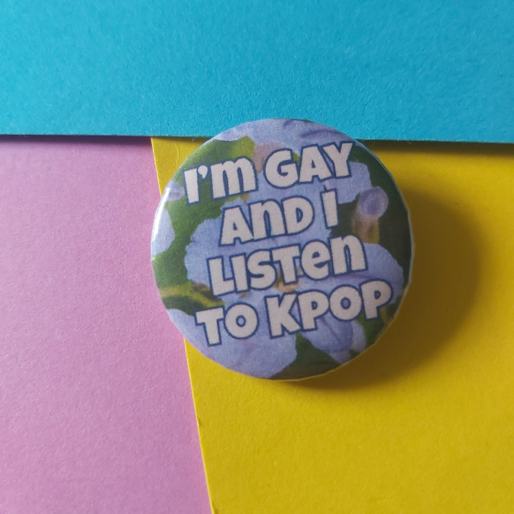 A round pinback button badge featuring the text 'I'm GAY AND I LISTEN TO KPOP' in white bubble letters over a background of light purple flowers and green leaves. The button is photographed on a background split diagonally into bright cyan-blue, yellow, and pink sections.