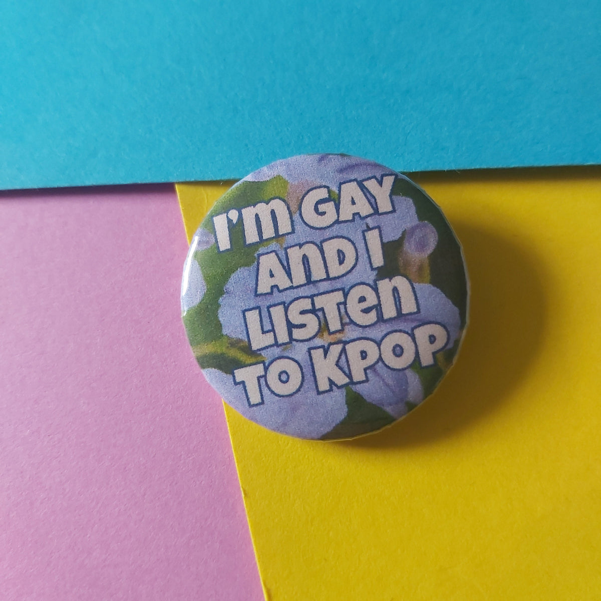 A round button pin with the text 'i'm GAY AND I LISTEN TO KPOP' in white lettering over a floral background, placed on a colorful background of blue, yellow, and pink paper.