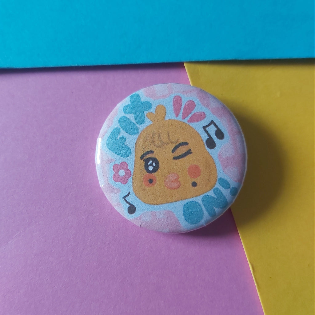 A round pin-back button badge featuring a cute cartoon chick character with the word 'FIX ON' and musical notes, displayed on a background of colorful paper blocks.