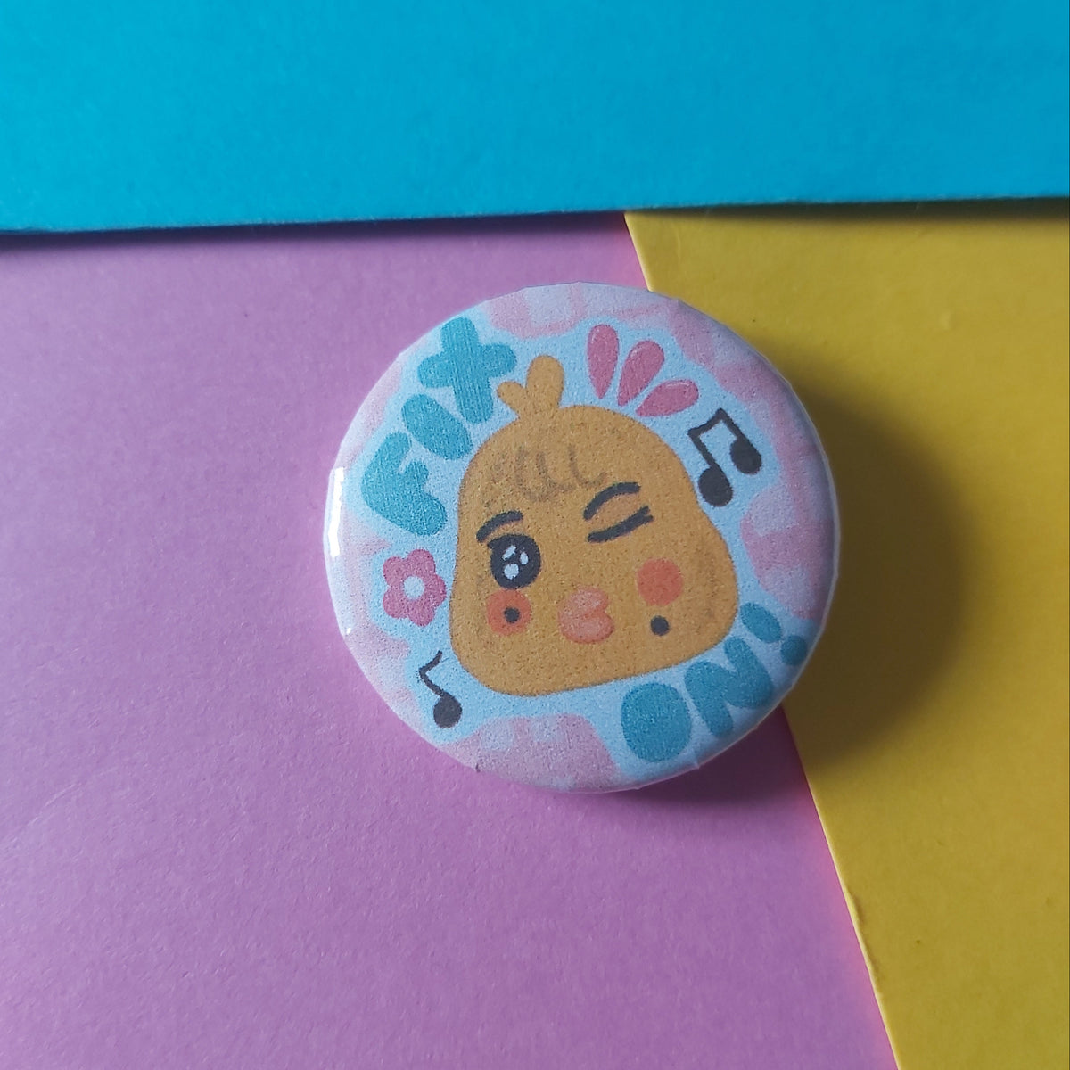 A round pin-back button badge featuring a cute cartoon chick character with the word 'FIX ON' and musical notes, displayed on a background of colorful paper blocks.