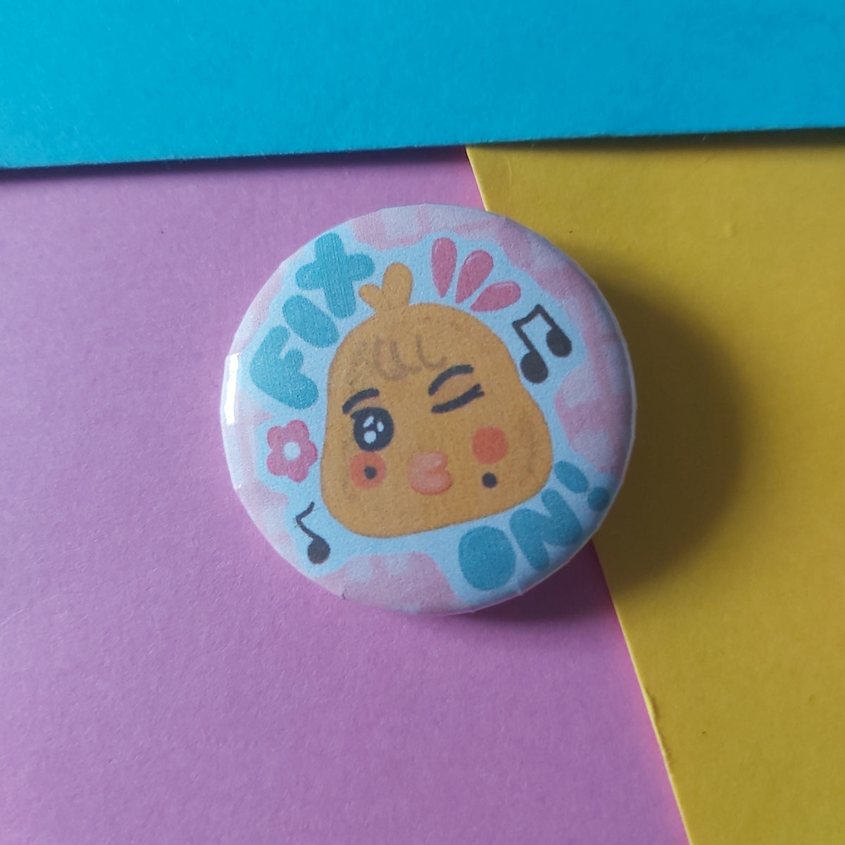 A round pin-back button badge featuring a cute chick cartoon character with musical notes and flowers, placed on a diagonally split background of yellow, purple, and blue paper.