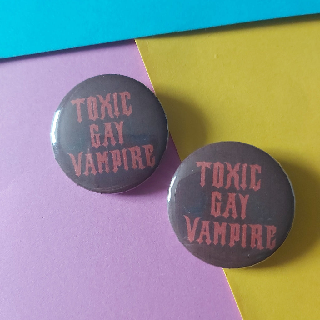 Two round pin-back buttons with a dark red background and pink text reading 'TOXIC GAY VAMPIRE', placed on a diagonally split yellow and pink paper background.