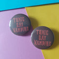 Two round pin-back buttons with a dark red background and pink text reading 'TOXIC GAY VAMPIRE', placed on a diagonally split yellow and pink paper background.