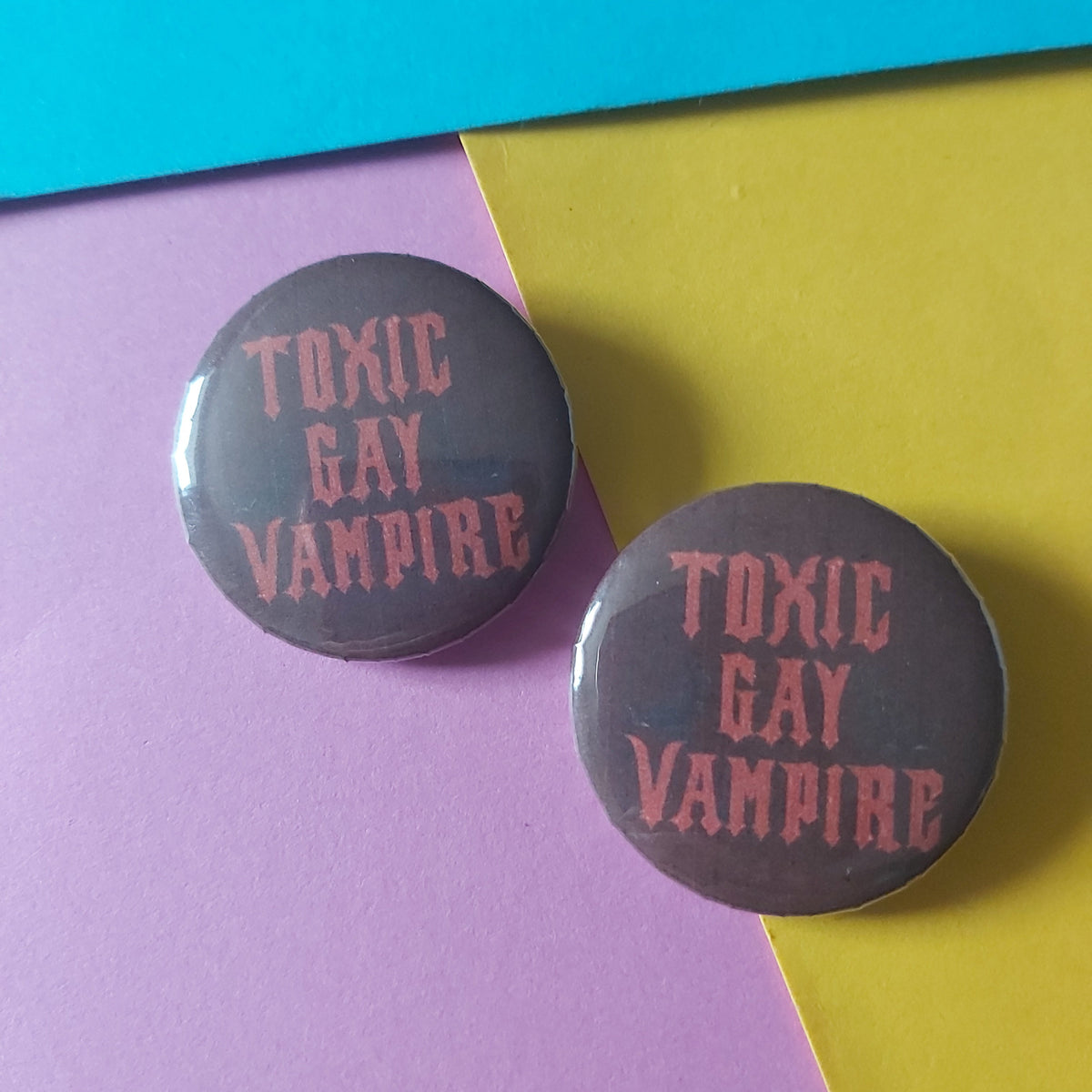 Two round pin-back buttons with a dark red background and pink text reading 'TOXIC GAY VAMPIRE', placed on a diagonally split yellow and pink paper background.