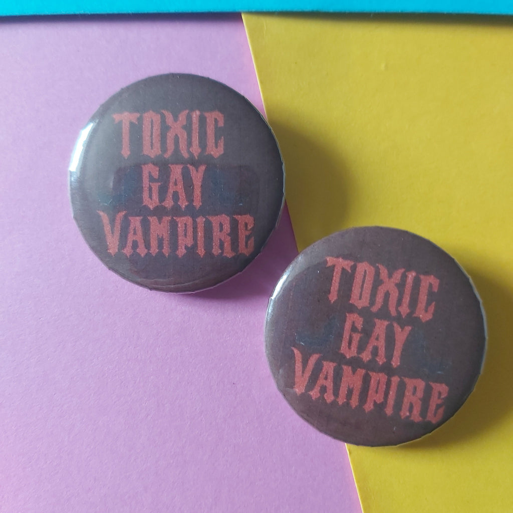 Two round pin-back buttons with the text 'TOXIC GAY VAMPIRE' in reddish pink on a dark red background, displayed on a diagonally split yellow and purple paper surface.