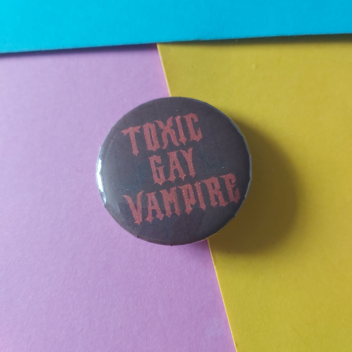 A round pin-back button badge with the text 'TOXIC GAY VAMPIRE' in a pinkish-red font on a dark red background, photographed on a split purple and yellow paper backdrop.