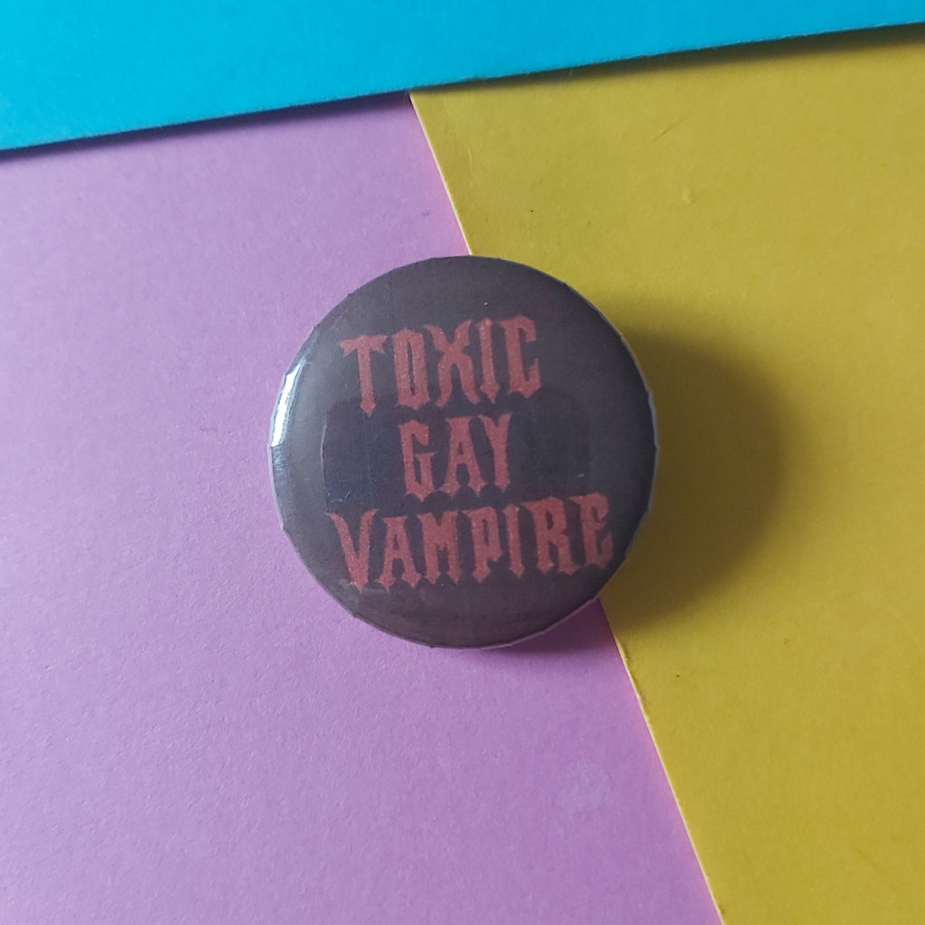 A round pinback button with the words 'TOXIC GAY VAMPIRE' in a red gothic font on a dark red background, photographed on a diagonally split yellow and pink surface.