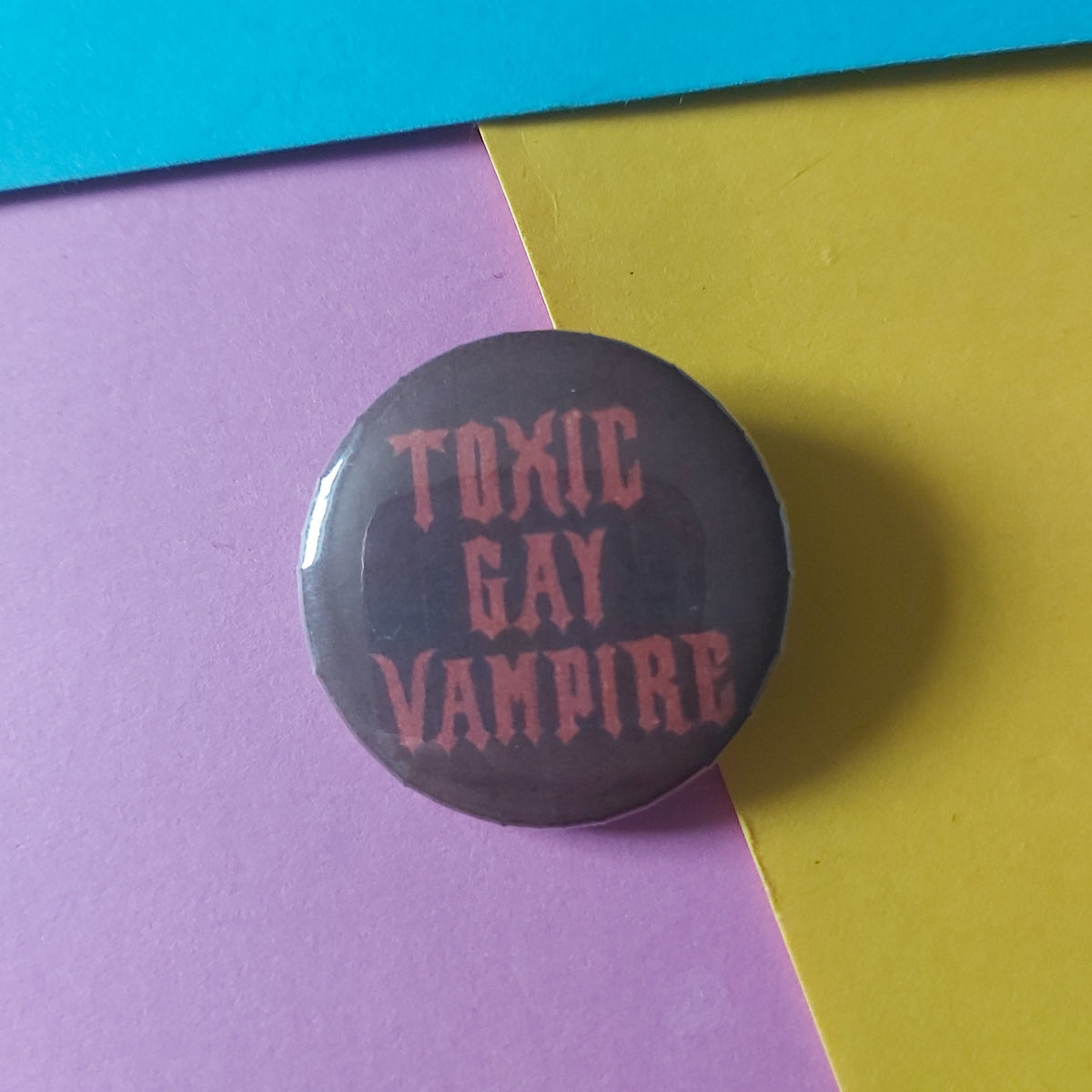A round pinback button badge featuring the text 'TOXIC GAY VAMPIRE' in a reddish-pink font on a dark red background, resting on a diagonally split yellow and pink surface with a blue strip on the left.