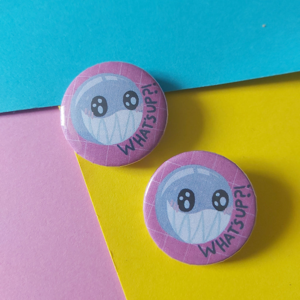 Two round pinback buttons featuring a round cute cartoon character with large eyes and the words 'WHAT'S UP?' in black text on a pink background, displayed on a colourful background of blue, yellow, and pink paper.