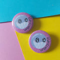 Two round pinback buttons featuring a round cute cartoon character with large eyes and the words 'WHAT'S UP?' in black text on a pink background, displayed on a colourful background of blue, yellow, and pink paper.