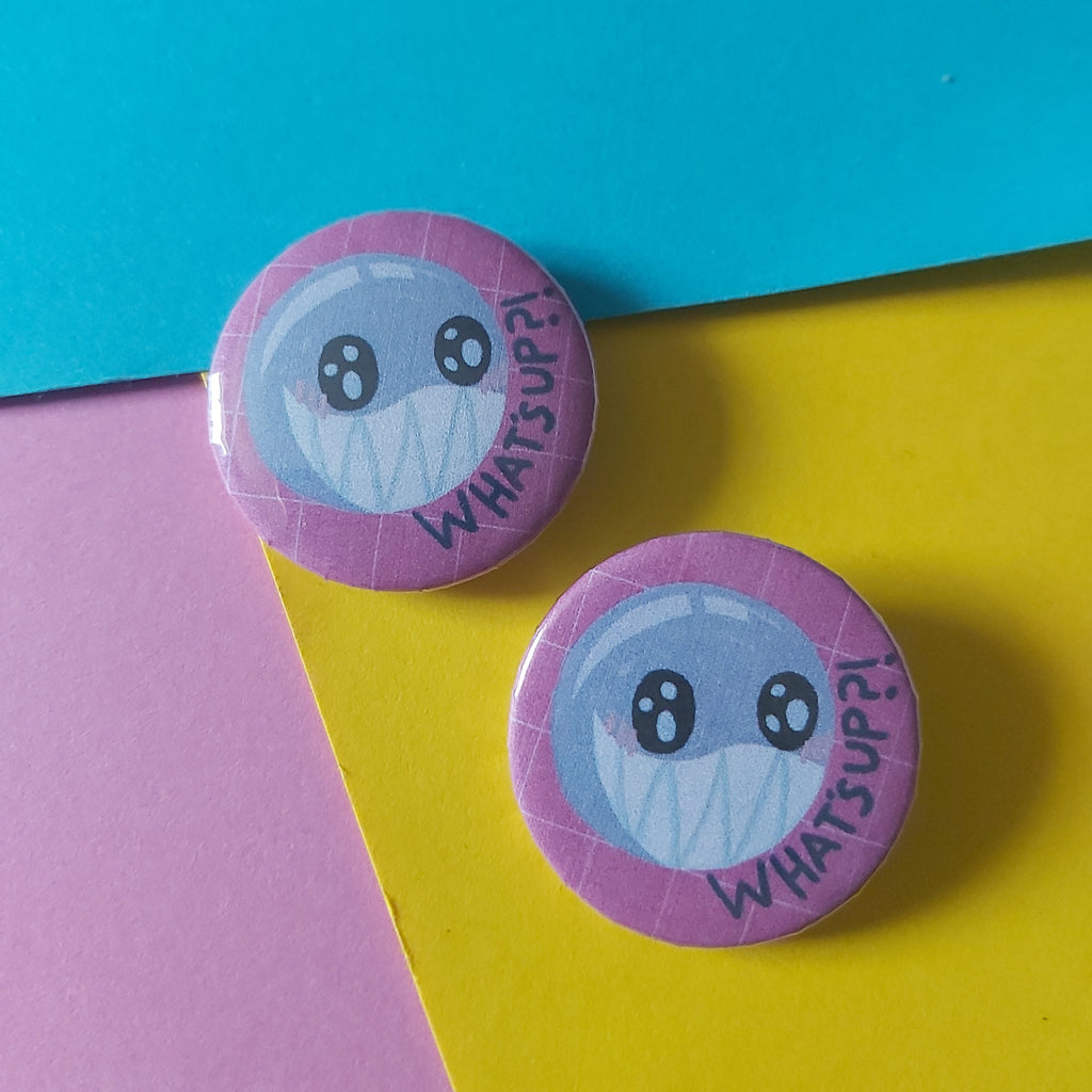 Two round pin-back buttons featuring a cartoon bubble character and the words 'WHAT'S UP?', displayed on a colourful background of cyan, yellow, and pink paper.