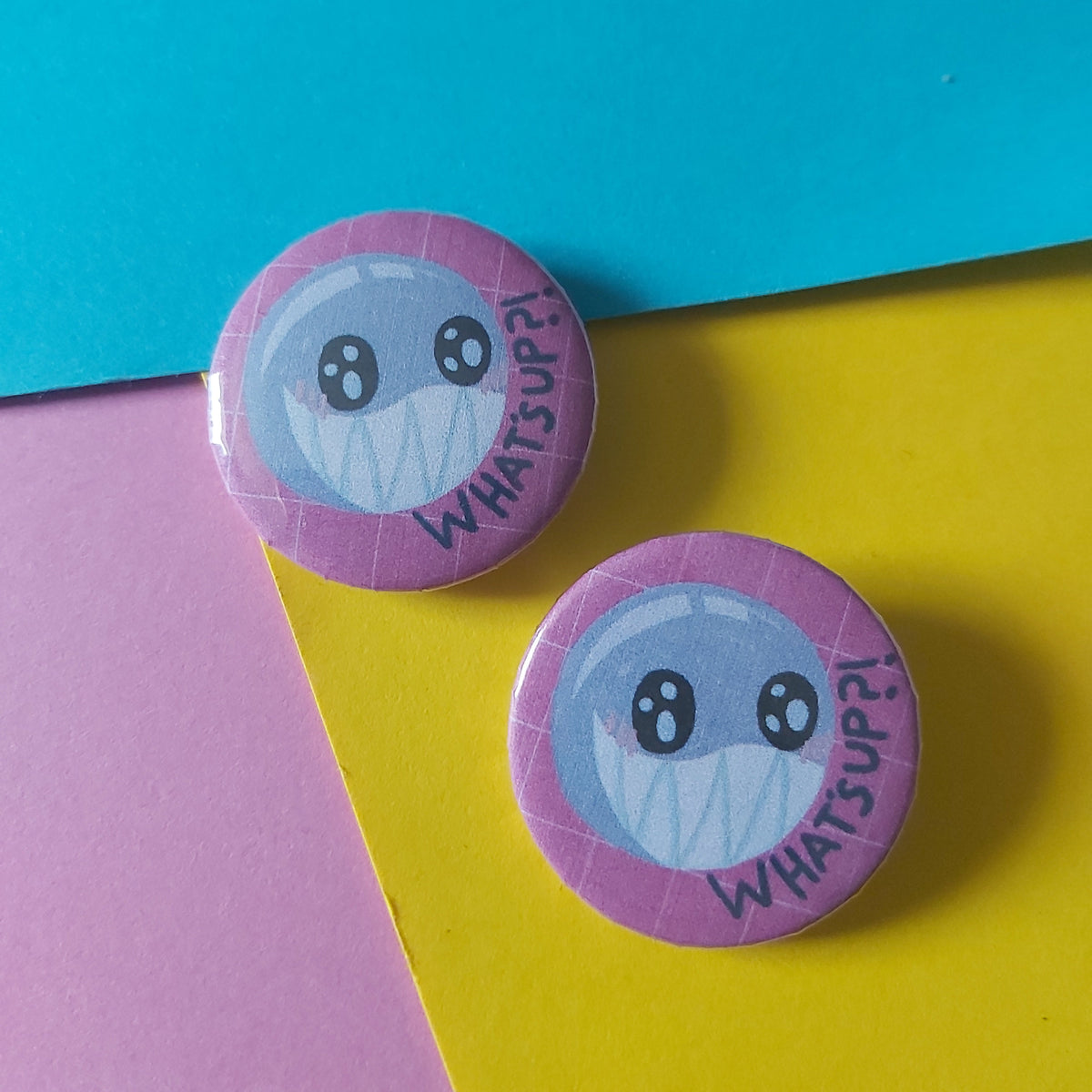 Two round pin-back buttons featuring a cartoon bubble character and the words 'WHAT'S UP?', displayed on a colourful background of cyan, yellow, and pink paper.