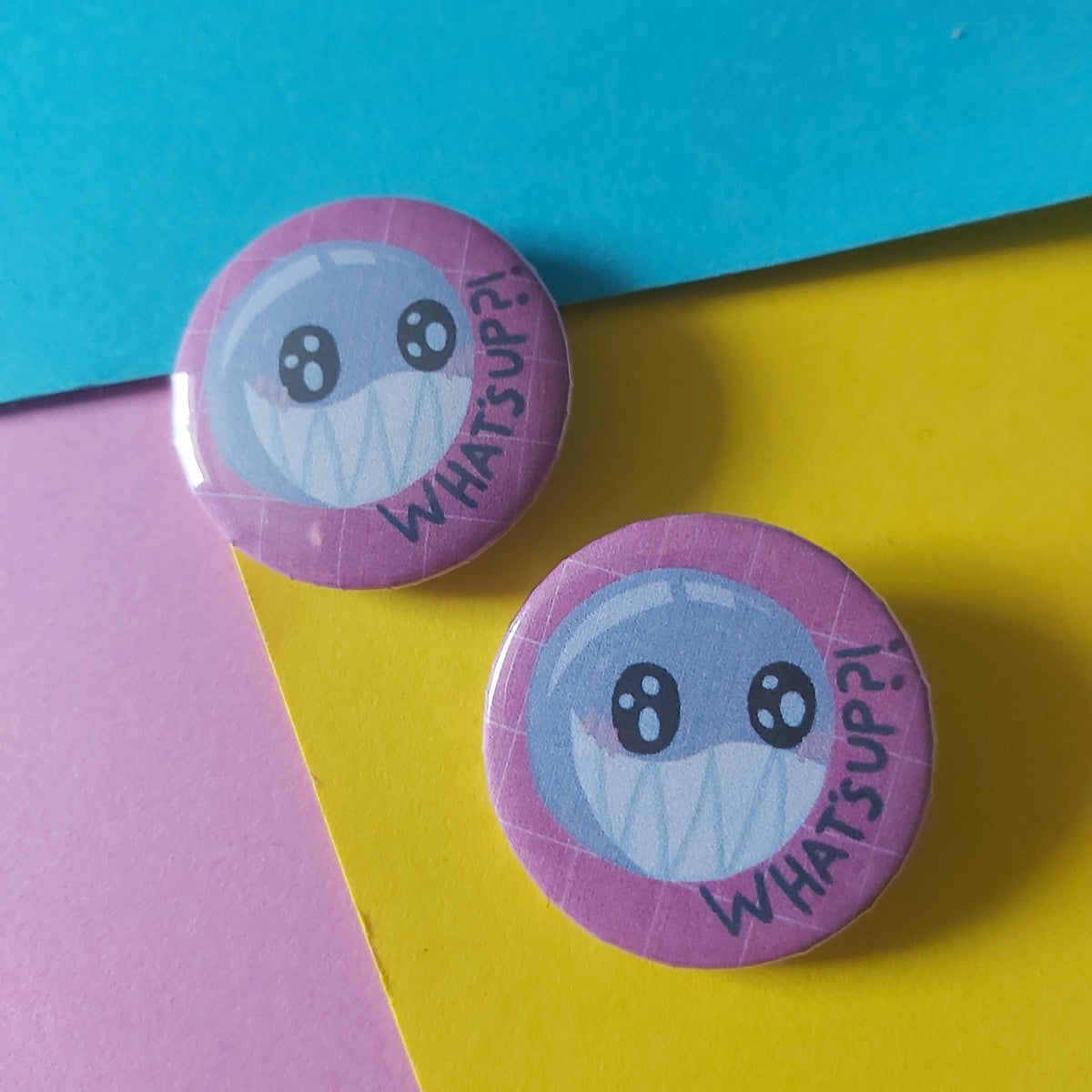 Two round pinback buttons featuring a cartoon bubble face with the words 'what's up?' on a pink background, displayed on a colourful background of blue, yellow, and pink paper.