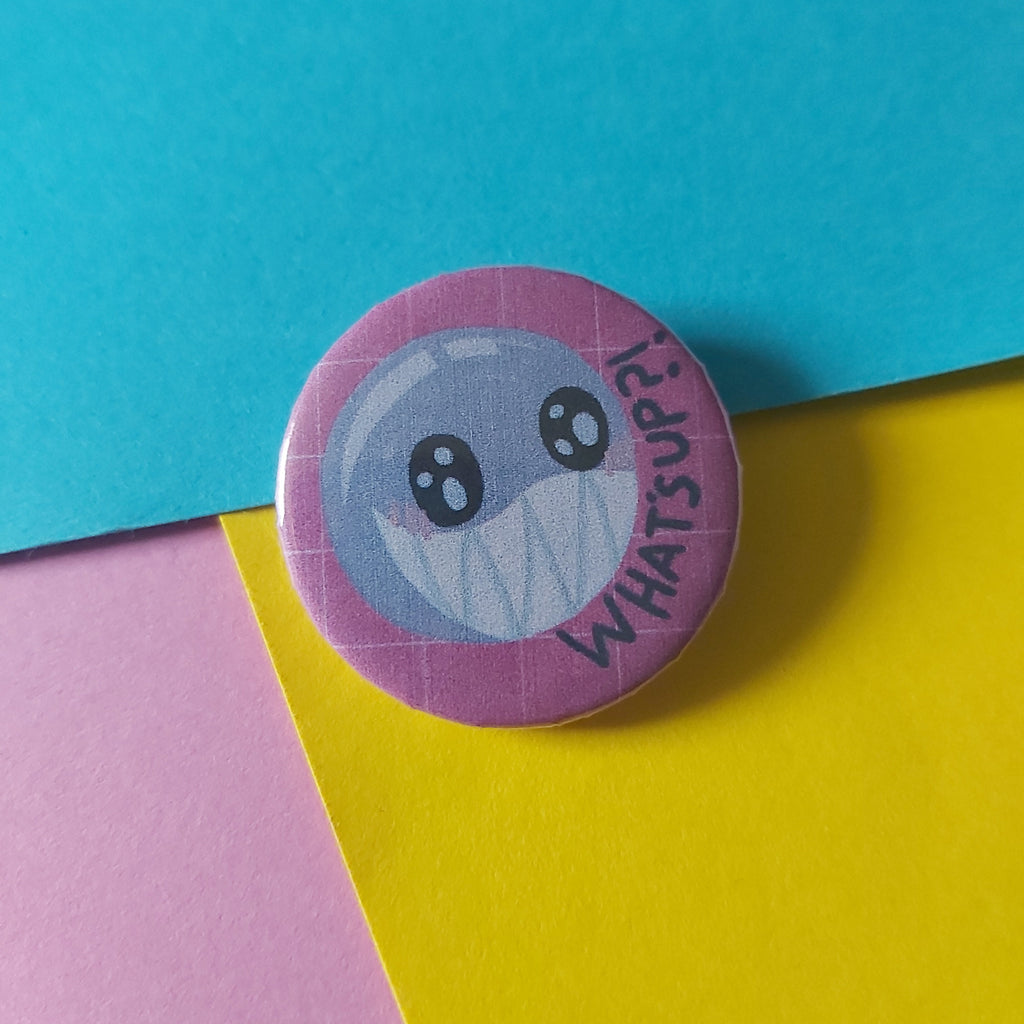 A round pinback button badge featuring a cartoon face with the text 'WHAT'S UP?' on a pink border, placed on a colorful background of blue, yellow, and pink paper.