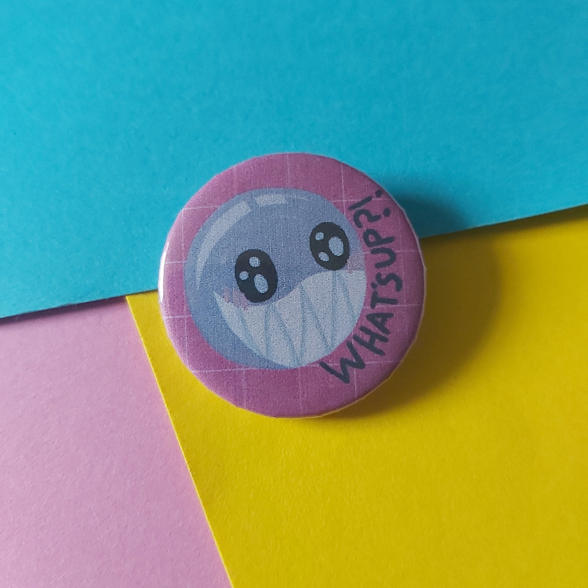 A round pinback button badge featuring a cartoon face with the text 'WHAT'S UP?' on a pink border, placed on a colorful background of blue, yellow, and pink paper.