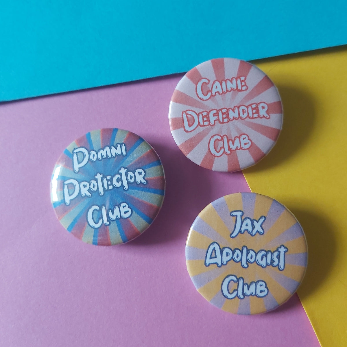Three round pin-back buttons with colourful sunburst designs and humorous club names ('Caine Defender Club', 'Jax Apologist Club', 'Pomni Protector Club') on a split yellow and purple background