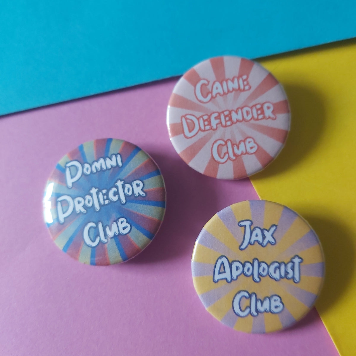 Three round pinback buttons with colourful sunburst designs and text reading 'Caine Defender Club,' 'Jax Apologist Club,' and 'Pomni Protector Club,' arranged on a split yellow and pink background with a blue corner.