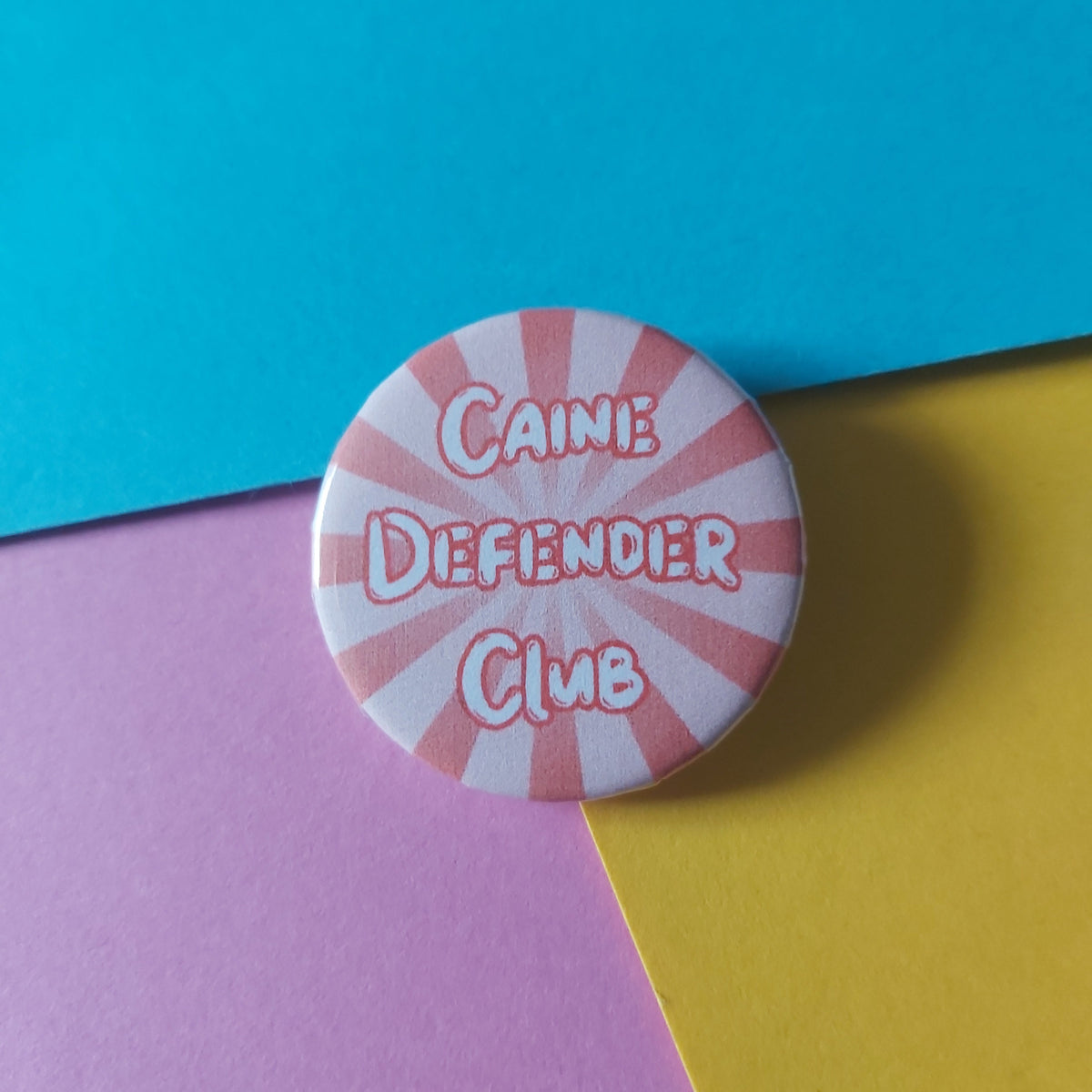 A round pin badge with a red and white sunburst design and the text 'CAINE DEFENDER CLUB' in stylised lettering, placed on a colourful background of blue, yellow, and pink paper.