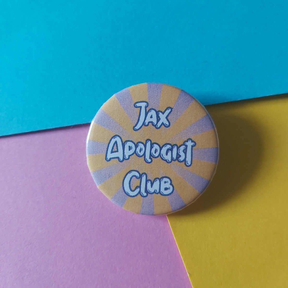 A round pin-back button badge with a yellow and lavender sunburst pattern and the text 'Jax Apologist Club' in blue, placed on a background of colored paper sections.