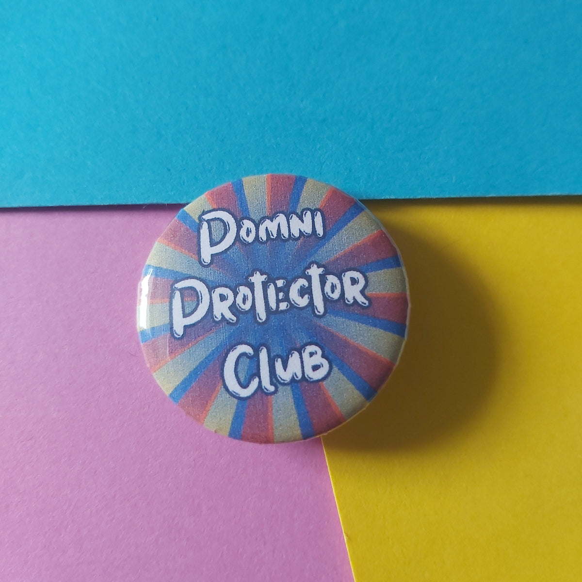 A round pin-back button badge with a colourful sunburst pattern in red, blue, and yellow, featuring the text 'Pomni Protector Club' in white. The badge is photographed on a background split diagonally between bright blue and yellow, with the bottom-right corner showing a pink section.