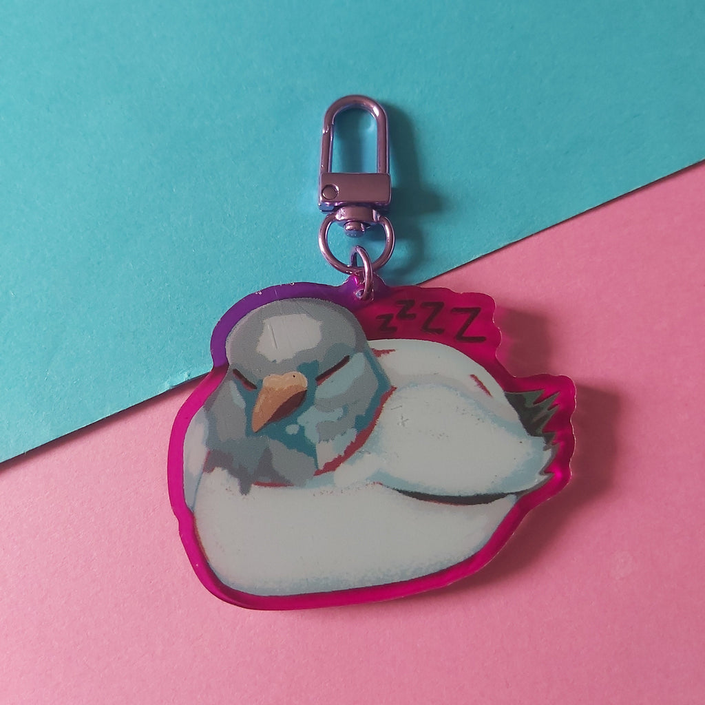 A die-cut acrylic keychain charm featuring a sleeping white bird with a pink purple outline, attached to a silver lobster clasp, set against a diagonally split pink and teal background.