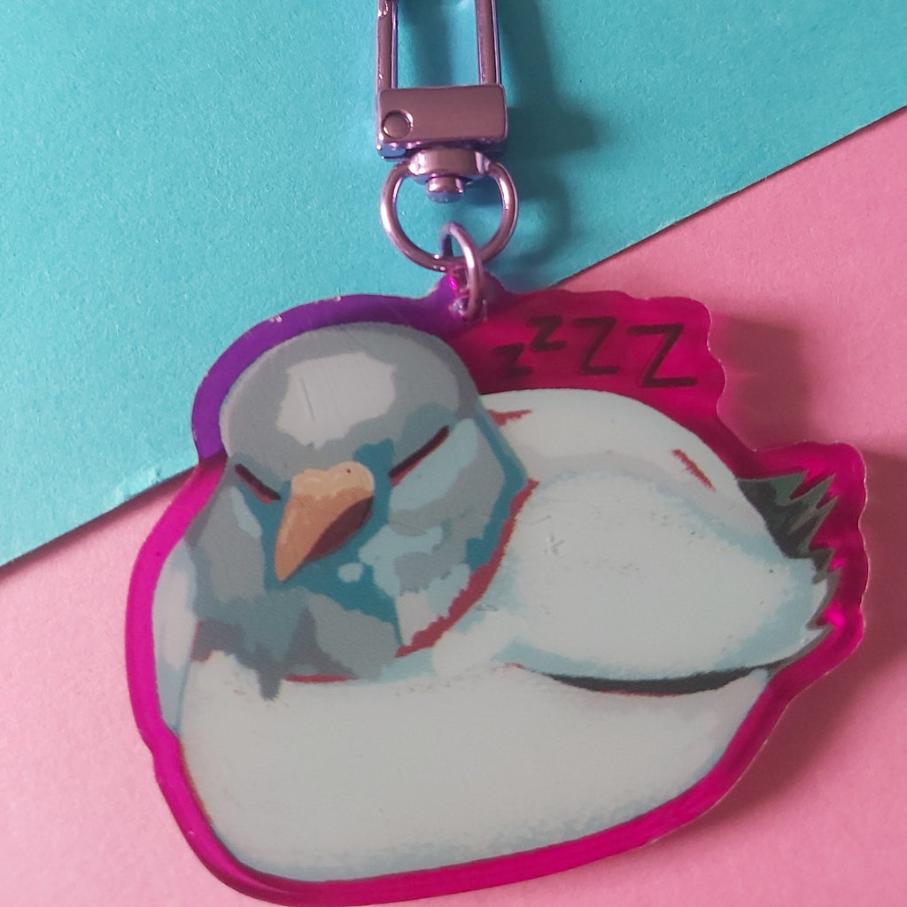 A sleeping white bird acrylic keychain charm with a pink-purple border, attached to a metallic pink clasp, set against a split turquoise and pink background.