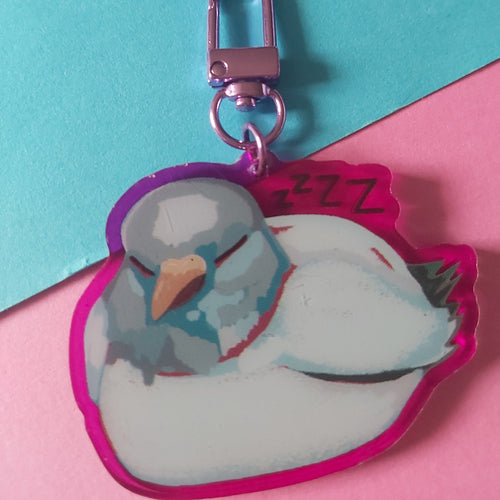 A sleeping white bird acrylic keychain charm with a pink-purple border, attached to a metallic pink clasp, set against a split turquoise and pink background.
