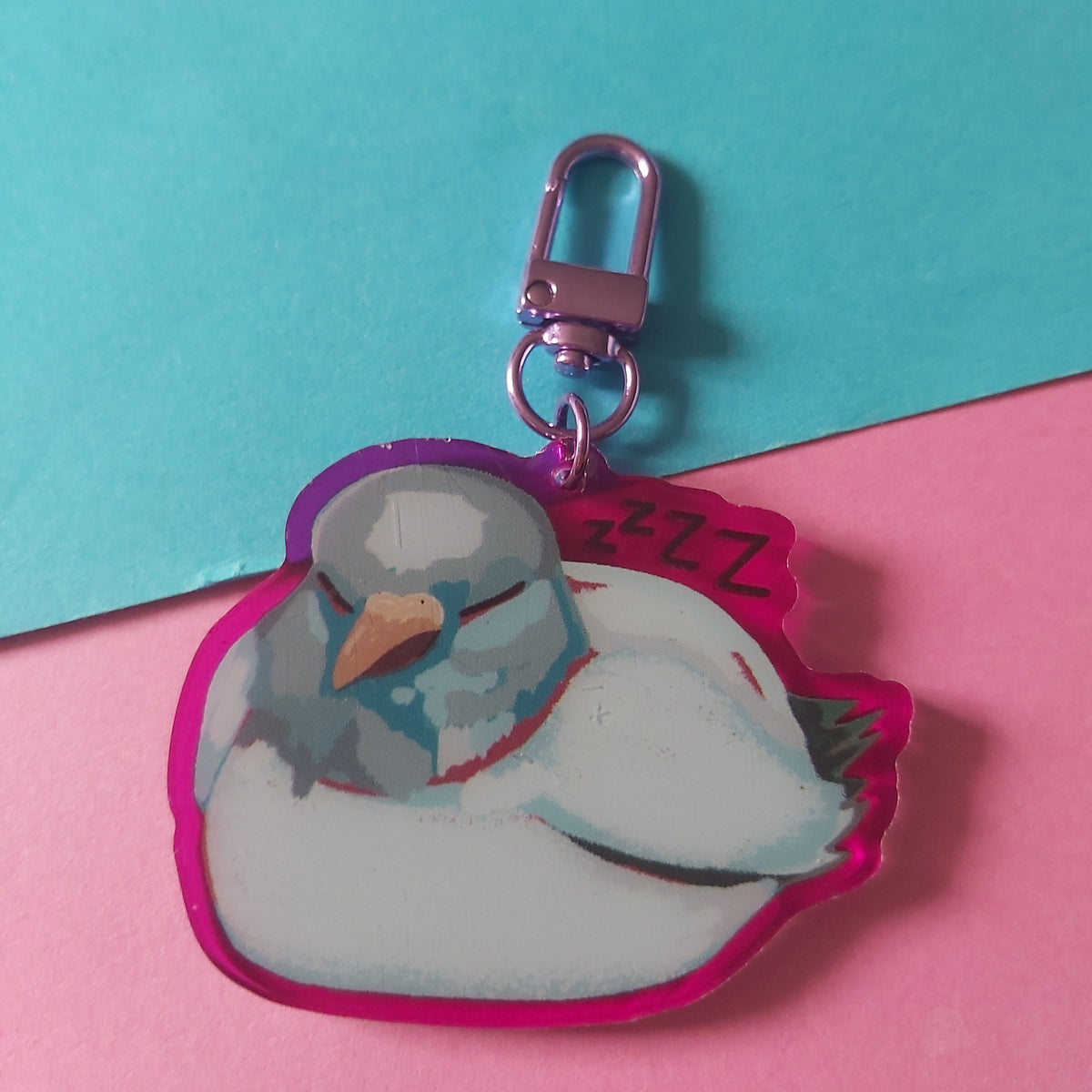 A die-cut acrylic keychain charm featuring a sleeping white bird with a pink-purple outline, attached to a metallic pink clasp, set against a split pink and teal background.