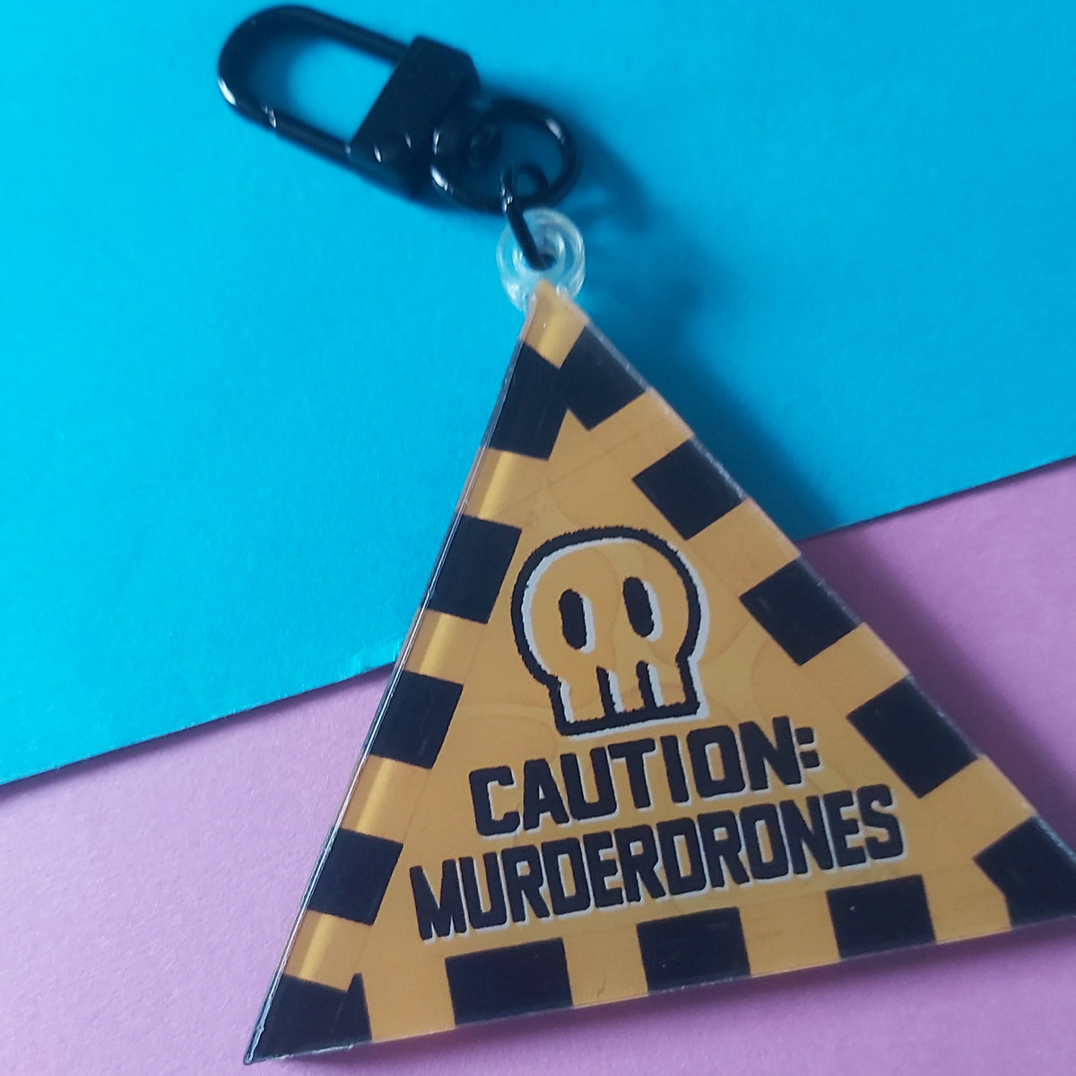 A triangular acrylic keychain featuring a yellow-and-black checkered pattern with a skull icon and the text 'CAUTION: MURDERDRONES'. The keychain is attached to a black carabiner clip and photographed on a split blue-and-pink background.