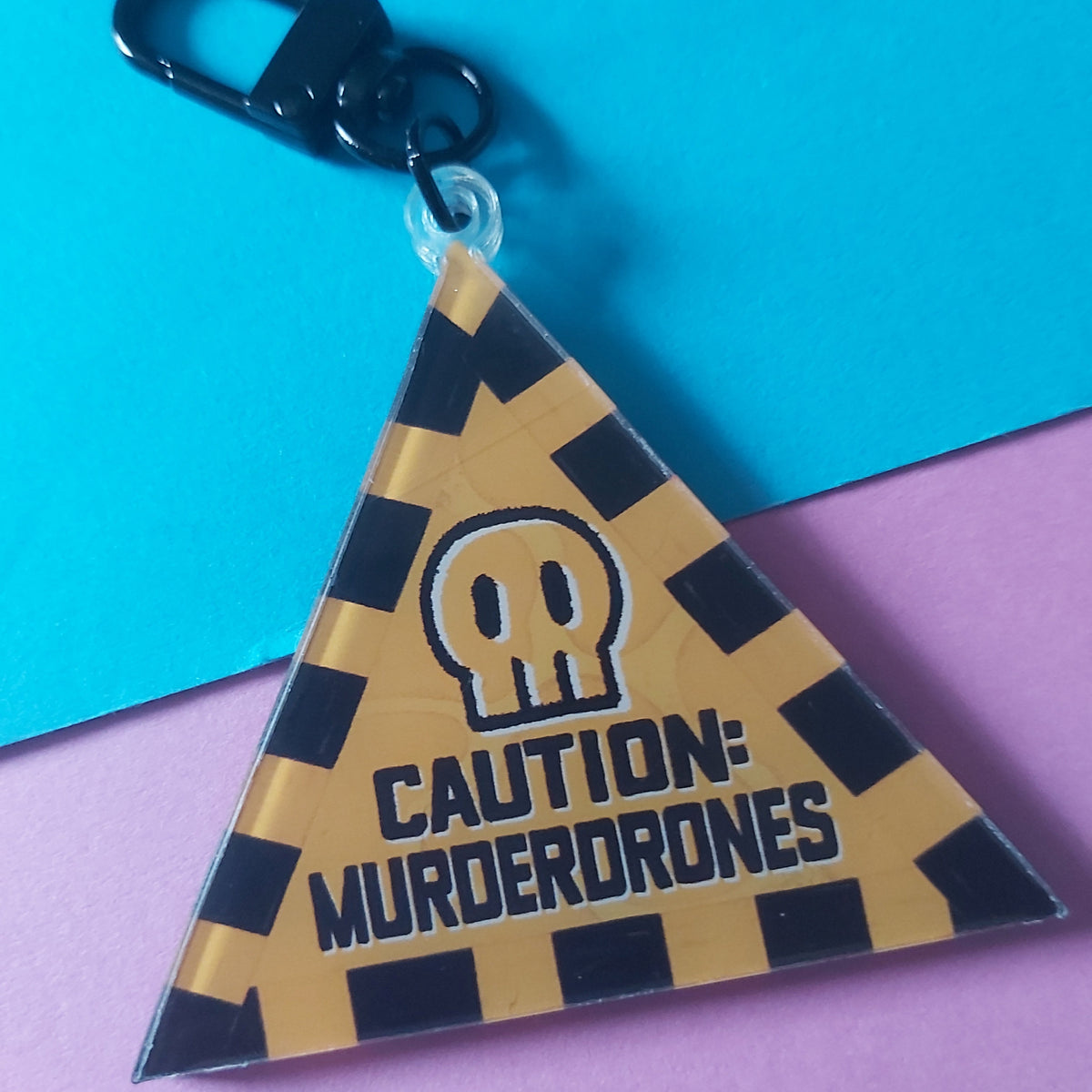 A triangular acrylic keychain with a black-and-yellow caution-style pattern, featuring a skull icon and the text 'CAUTION: MURDERDRONES' in black. The keychain includes a black metal clasp and is photographed on a split blue and pink background.