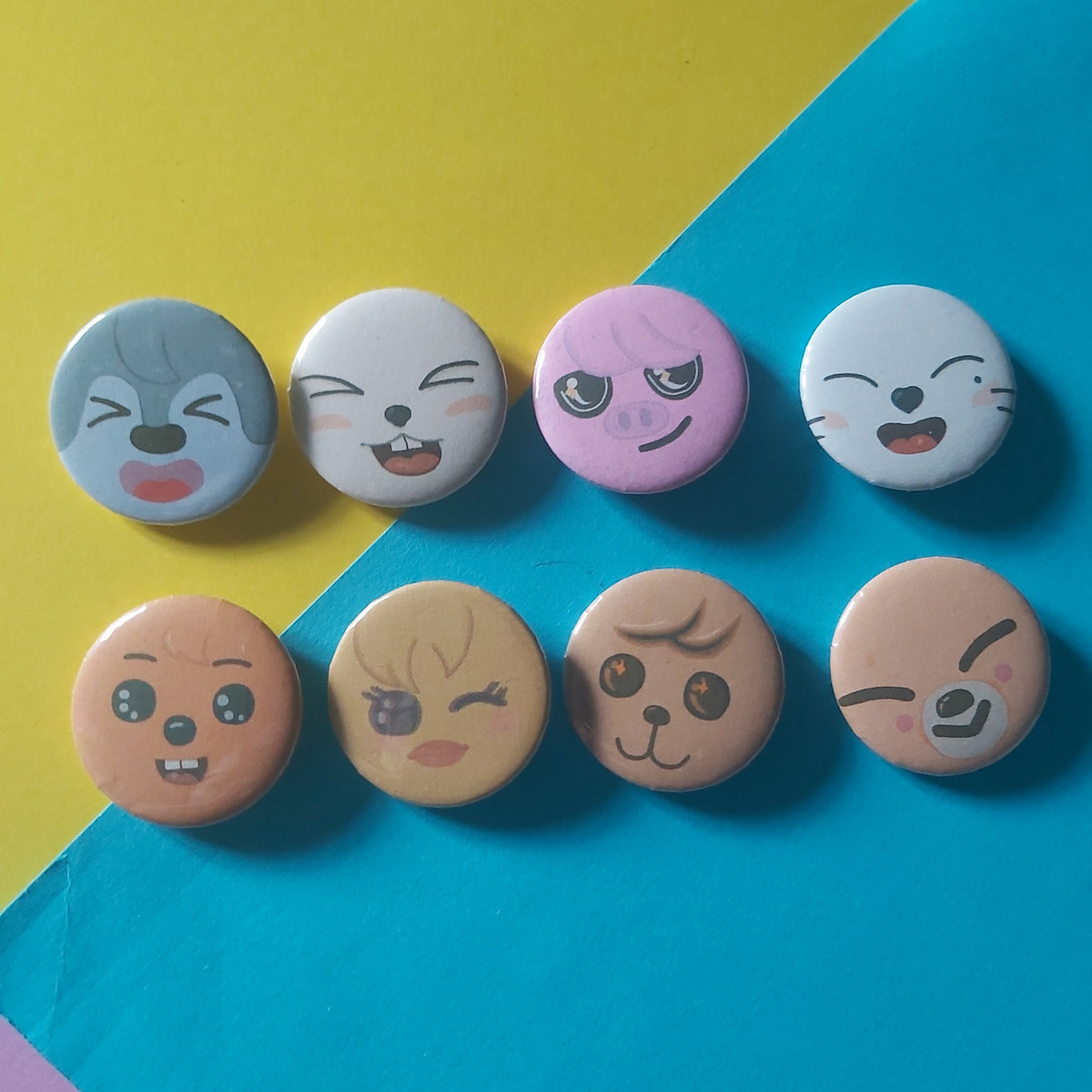 Eight round pinback buttons featuring cute cartoon faces in various expressions, arranged in two columns on a diagonally split yellow and blue background.