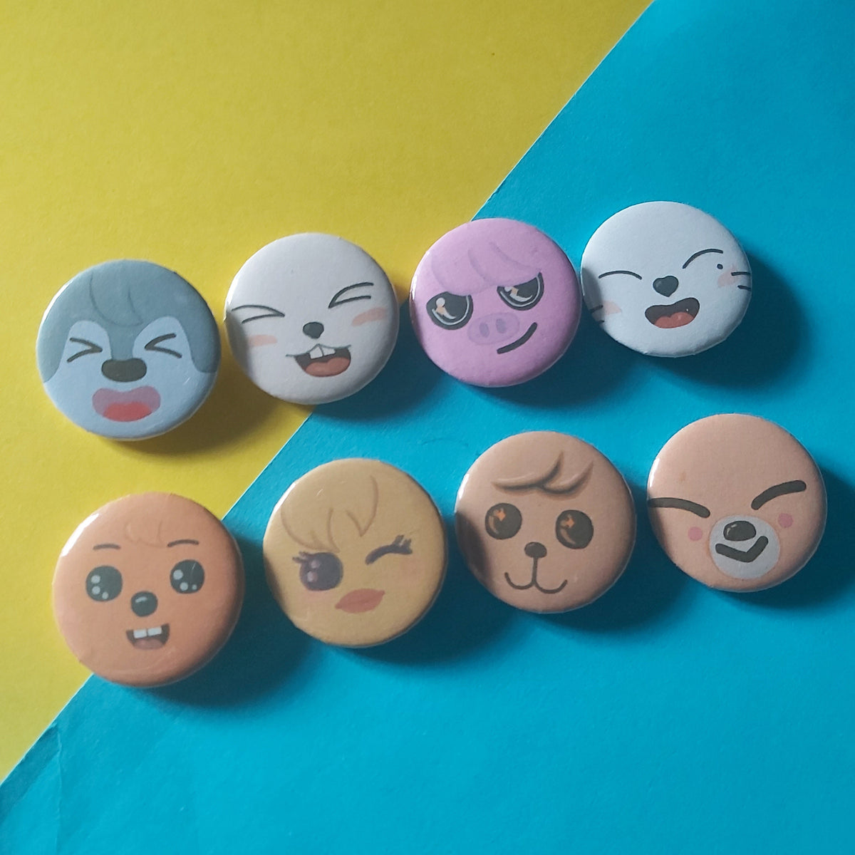 Eight small round pin-back buttons with cartoon face designs arranged in two vertical columns on a diagonally split yellow and bright blue background.