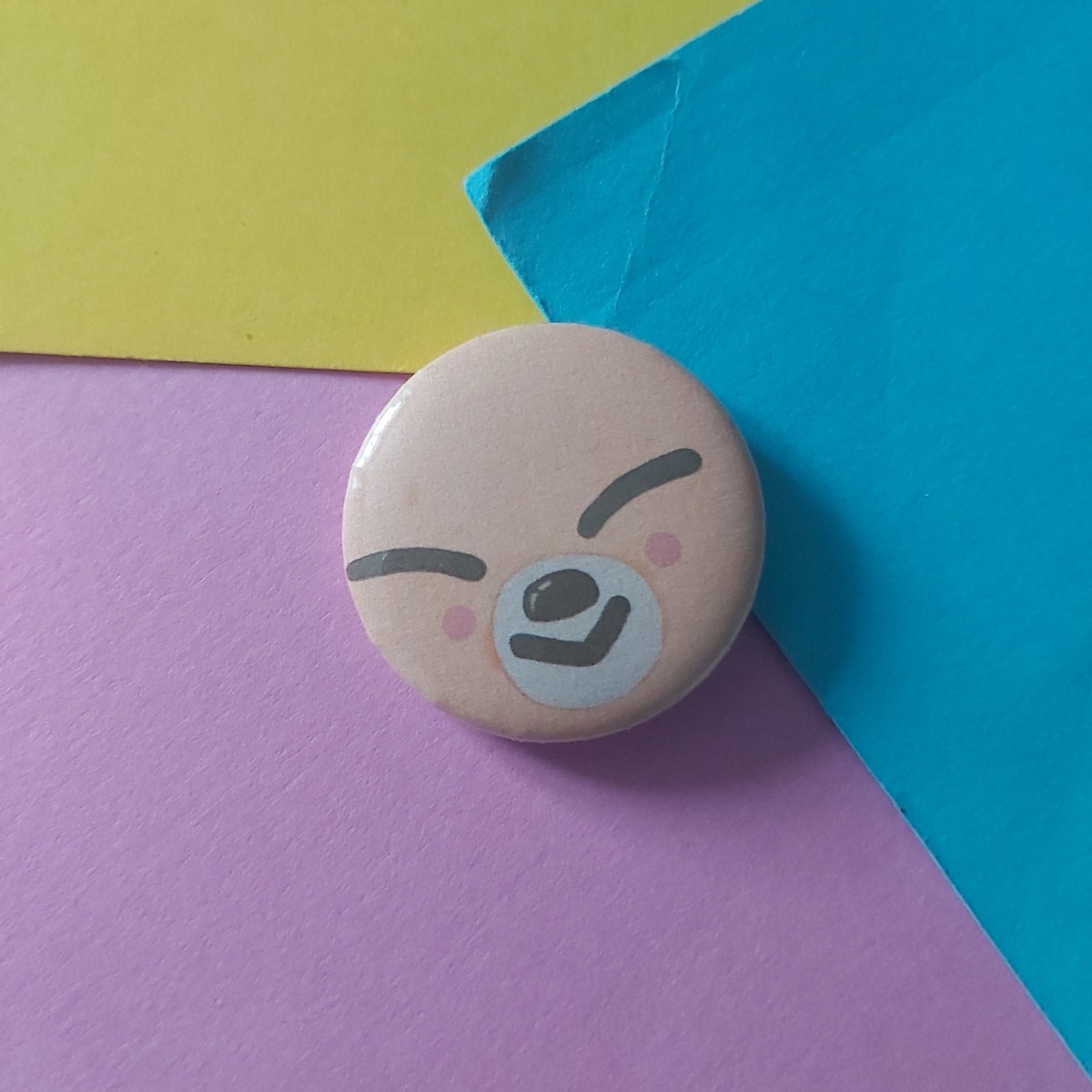 A small round pin badge featuring a cute cartoon fox face with closed eyes and pink cheeks, resting on a colourful background of yellow, blue, and pink paper.