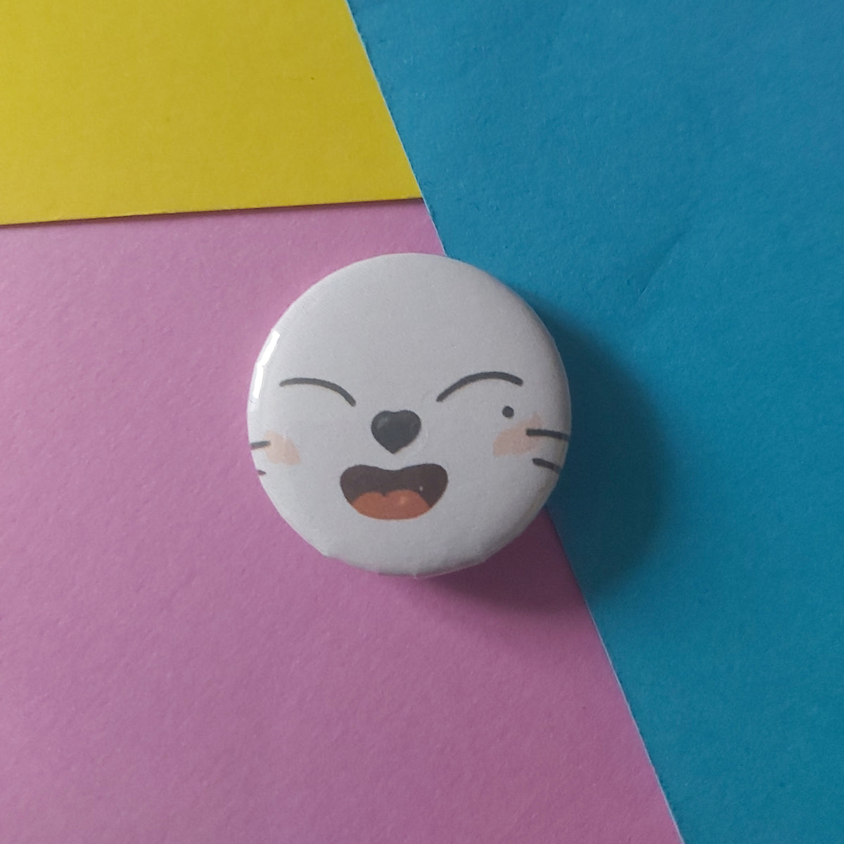 A round white button badge featuring a cartoon ferret face with closed eyes and an open mouth, placed on a background of blue, pink, and yellow paper.