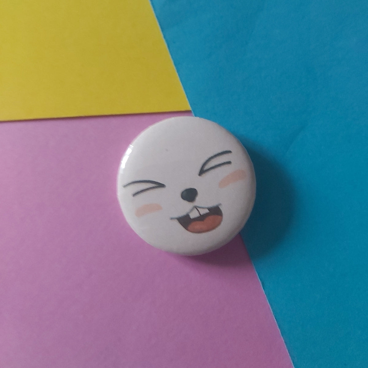A round white button pin featuring a cartoon rabbit face with closed eyes, resting on a colourful background of blue, yellow, and pink paper.