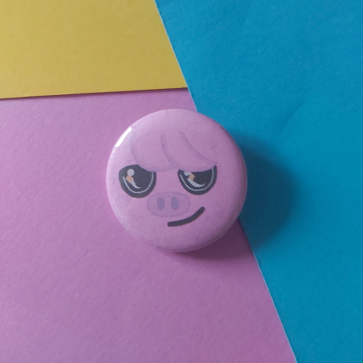 A round pink button badge featuring a cartoon pig face, placed on a background of diagonally split blue, yellow, and pink paper.