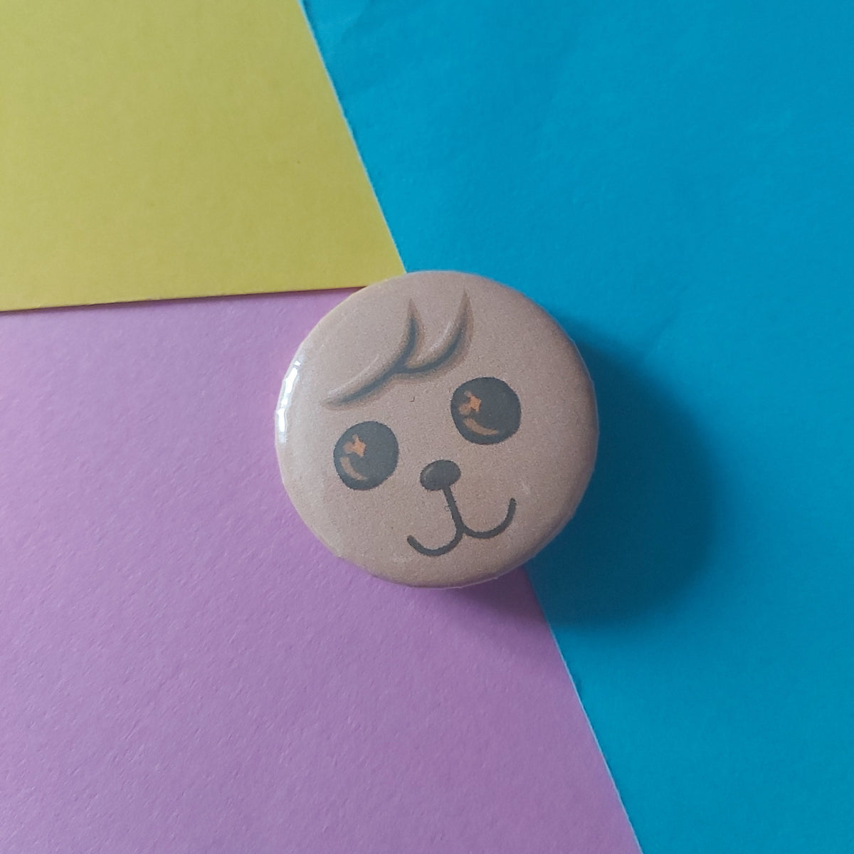 A round pin badge featuring a cute cartoon puppy face, resting on a diagonally split background of bright blue, yellow, and pink paper.