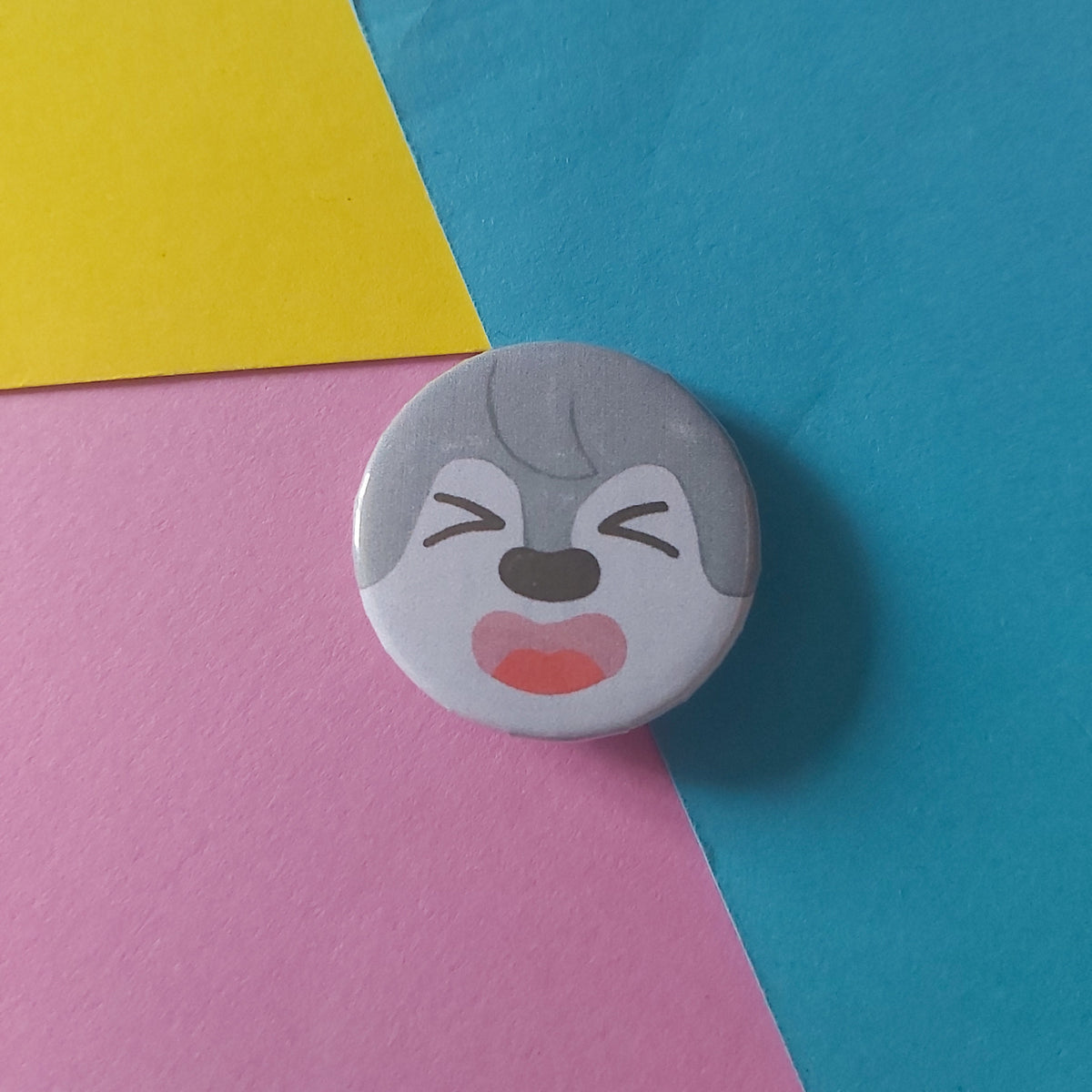 A round pin-back button badge featuring a cartoon wolf face with grey fur, closed eyes, and an open mouth, resting on a background of colourful paper in blue, yellow, and pink.