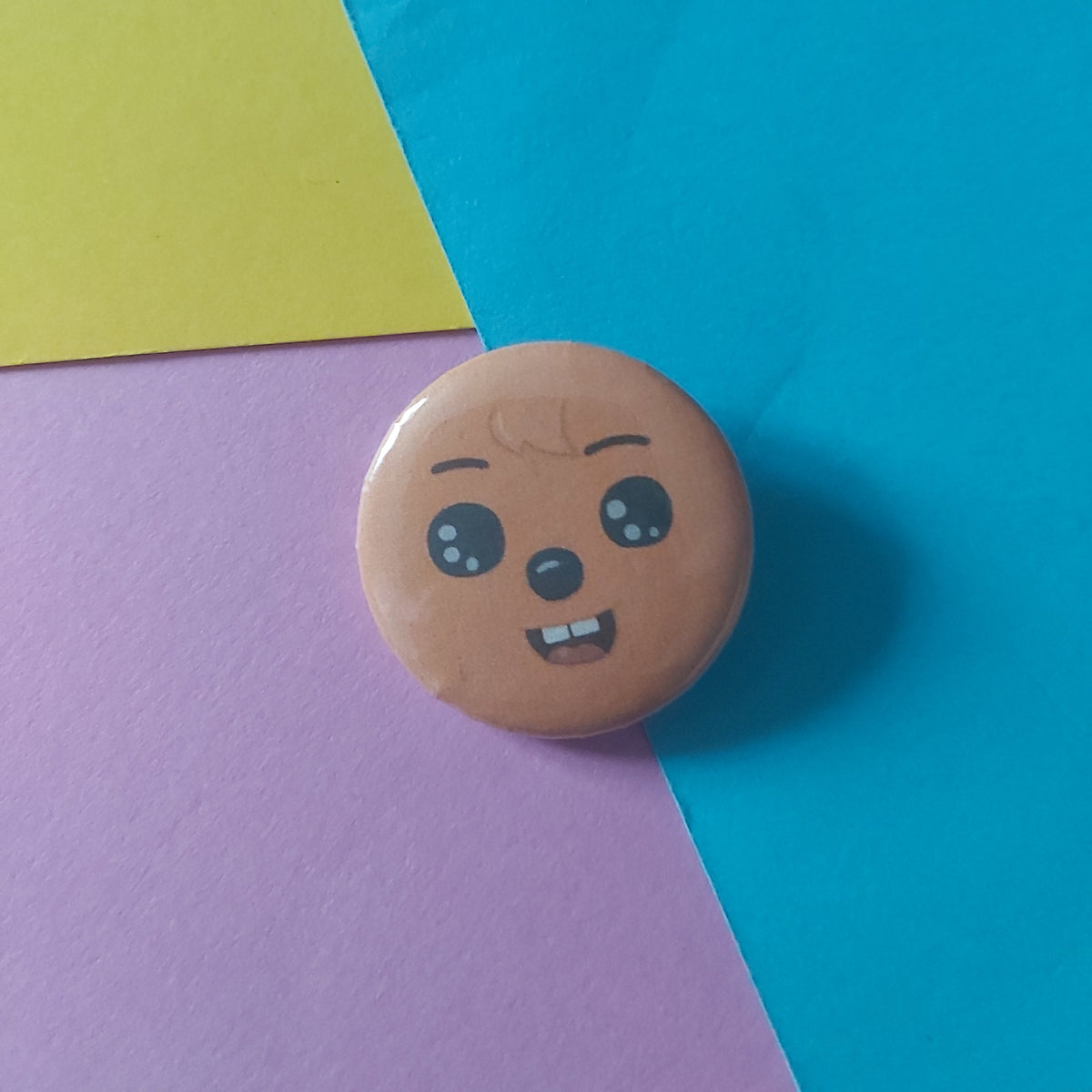 A round pin badge featuring a cartoon face quokka with brown skin and black eyes, resting on a geometric background of blue, pink, and yellow paper.